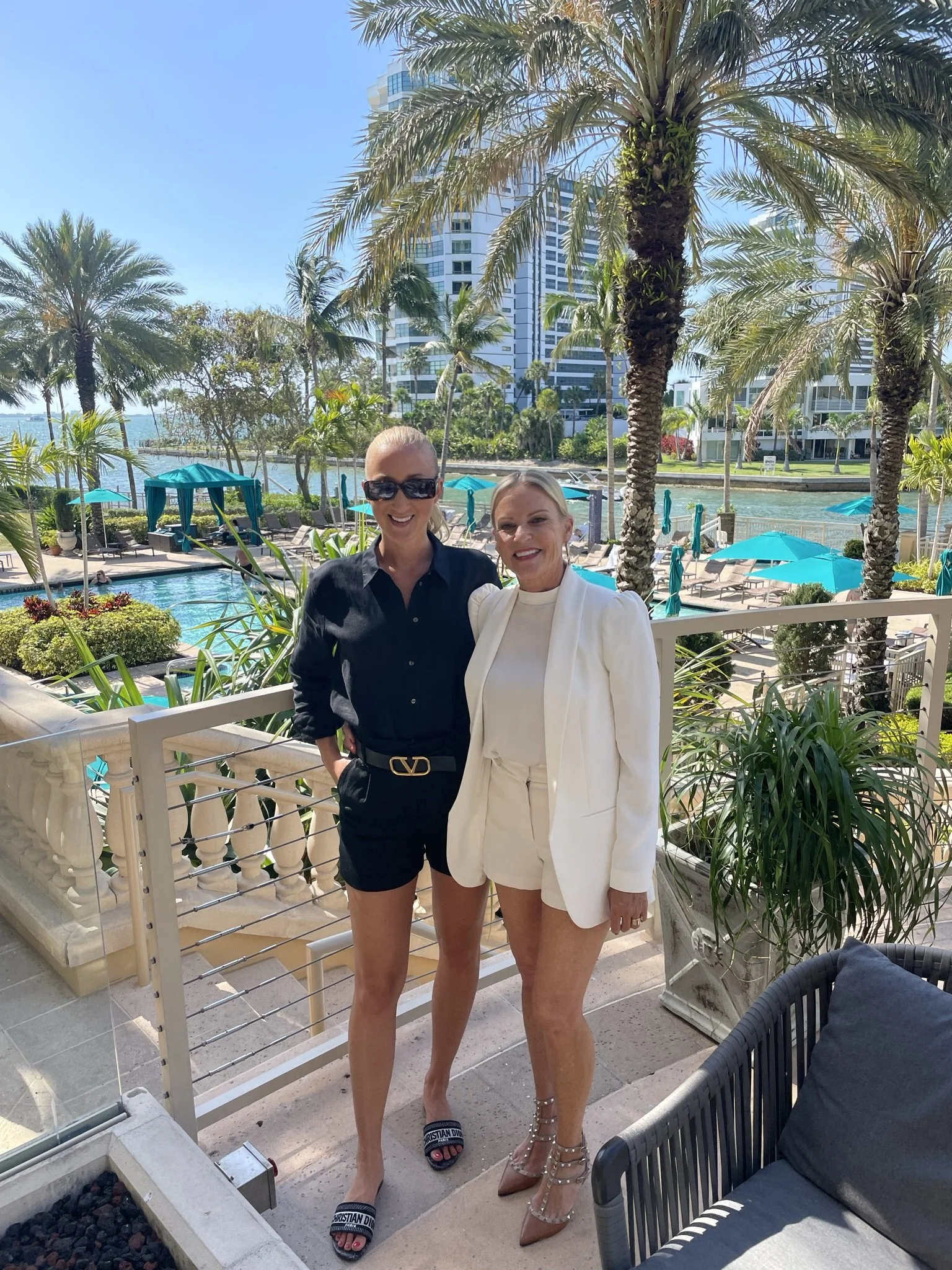 Two women standing on a balcony overlooking a landscaped pool area with palm trees, lounge chairs, umbrellas, and a waterway with high-rise buildings in the background. One woman is dressed in black with sunglasses, and the other in white with high heels.