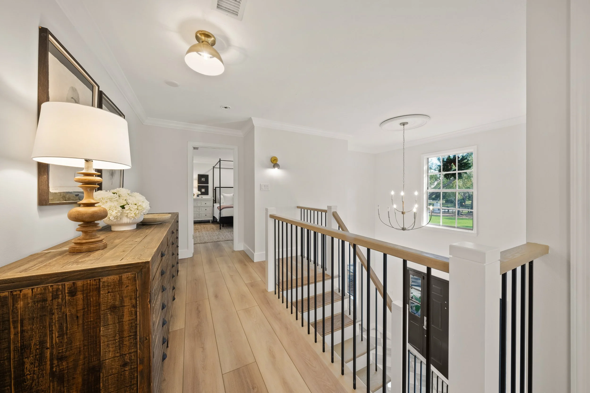 Bright hallway leading to a bedroom with a bed and white nightstands, featuring a staircase with dark metal railings and a chandelier, with a large window providing natural light.