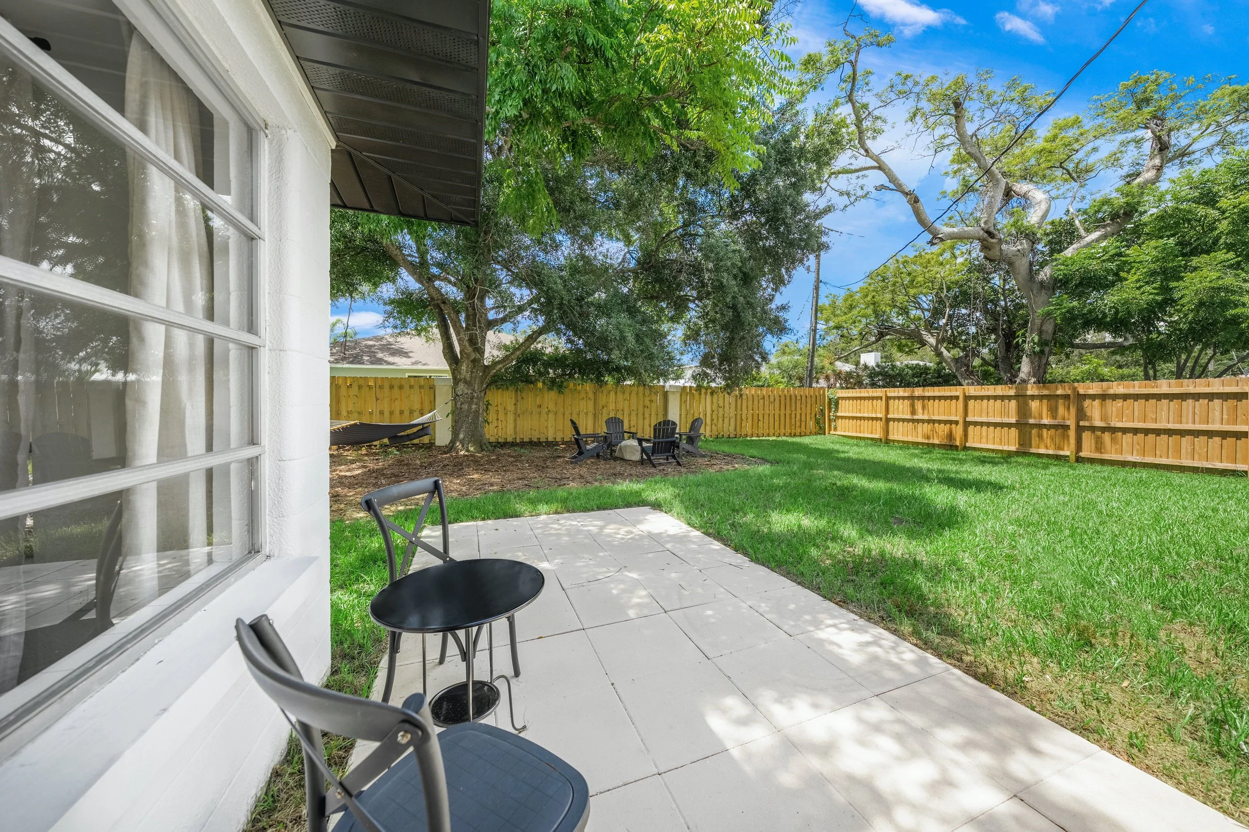 View of a backyard patio with two black chairs and a small black table, green grass, a large tree providing shade, a wooden fence, and additional seating in the distance under another tree; part of the house with a window in the foreground.