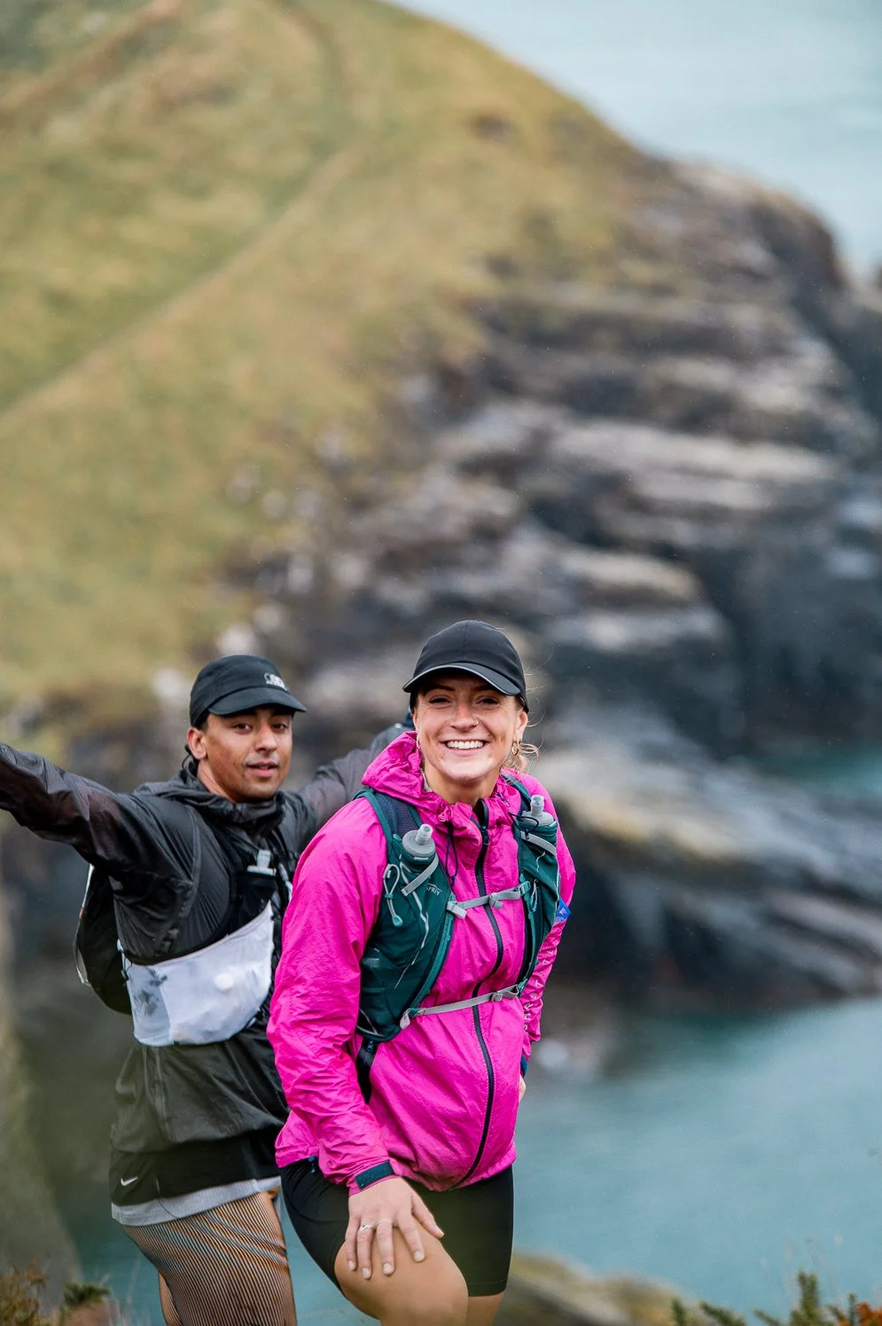 Two hikers, a man and a woman, smiling outdoors near water with rocky cliffs in the background.
