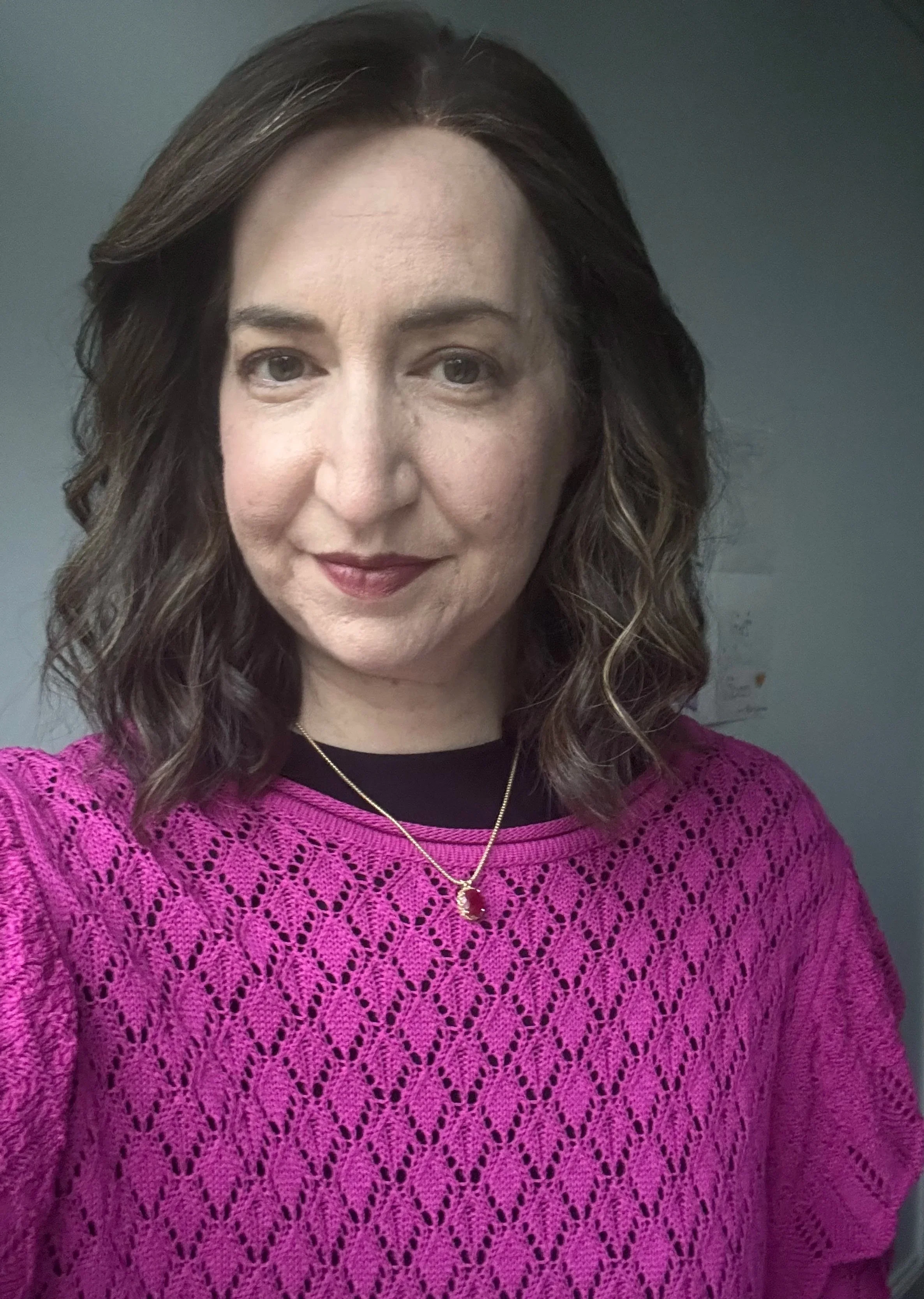 A woman with shoulder-length brown wavy hair, wearing a black top underneath a bright pink crochet sweater and a gold necklace with a pink pendant, smiling slightly at the camera against a plain gray background.