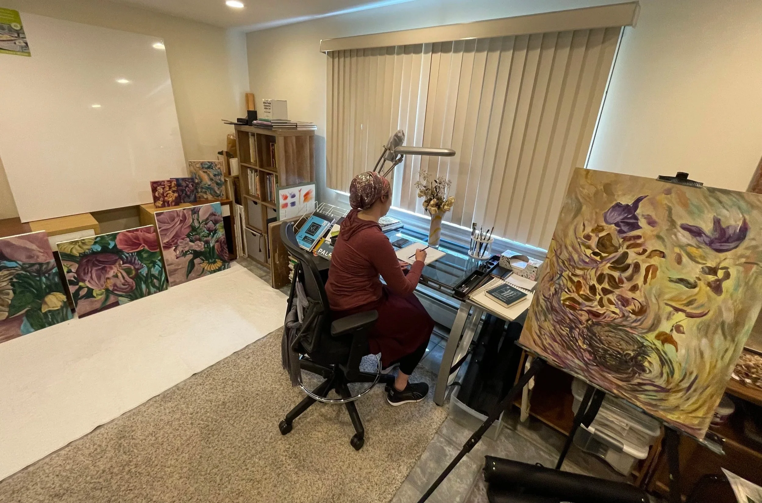 A person in a maroon dress, sitting at a glass desk in an art studio. They are working on a sketchbook with art supplies nearby. The studio has several finished paintings leaning against the wall, an easel with a colorful abstract painting, and a window with closed vertical blinds.