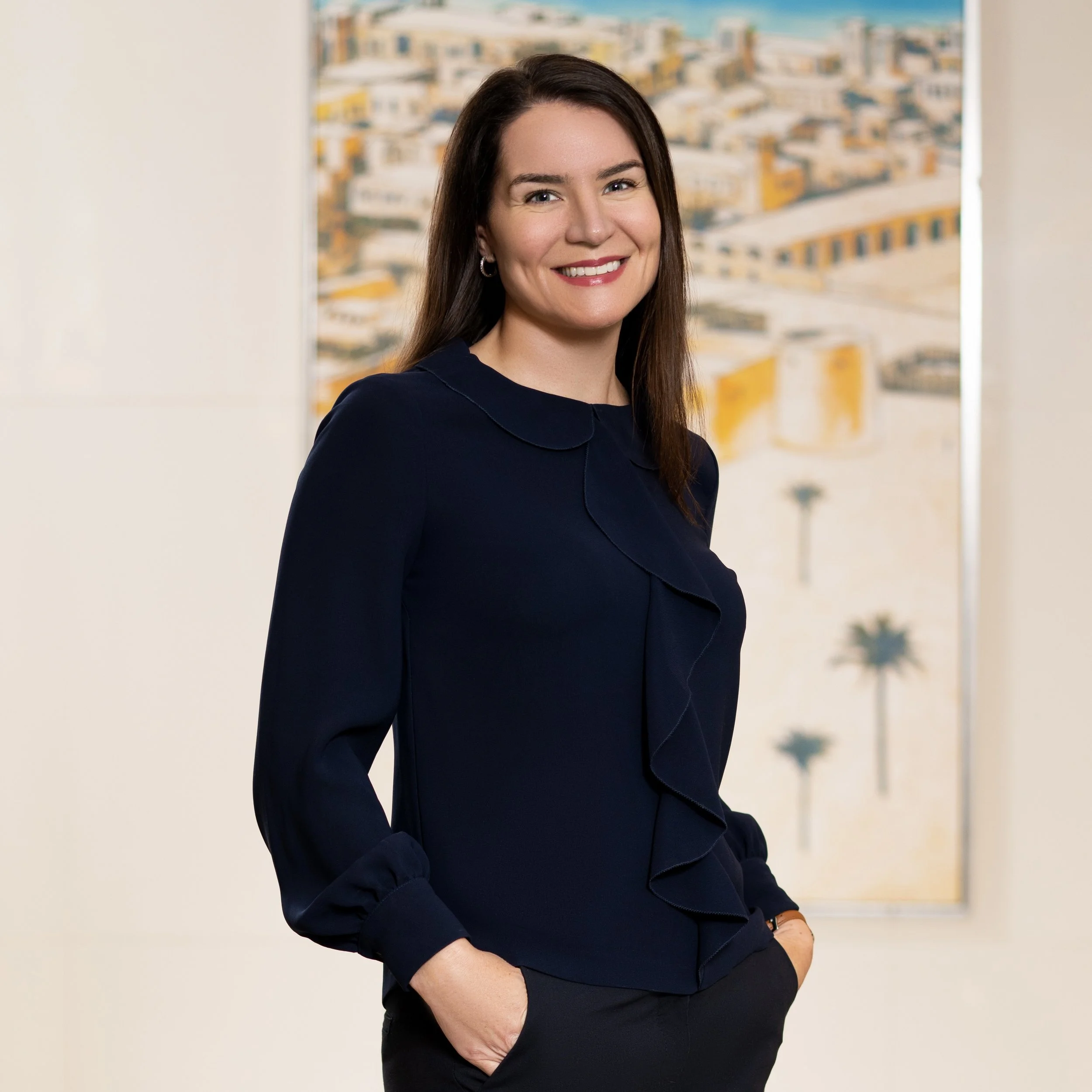 A woman with long dark brown hair smiling, wearing a navy blue blouse with ruffles and black pants, standing in front of abstract art depicting a cityscape with palm trees.