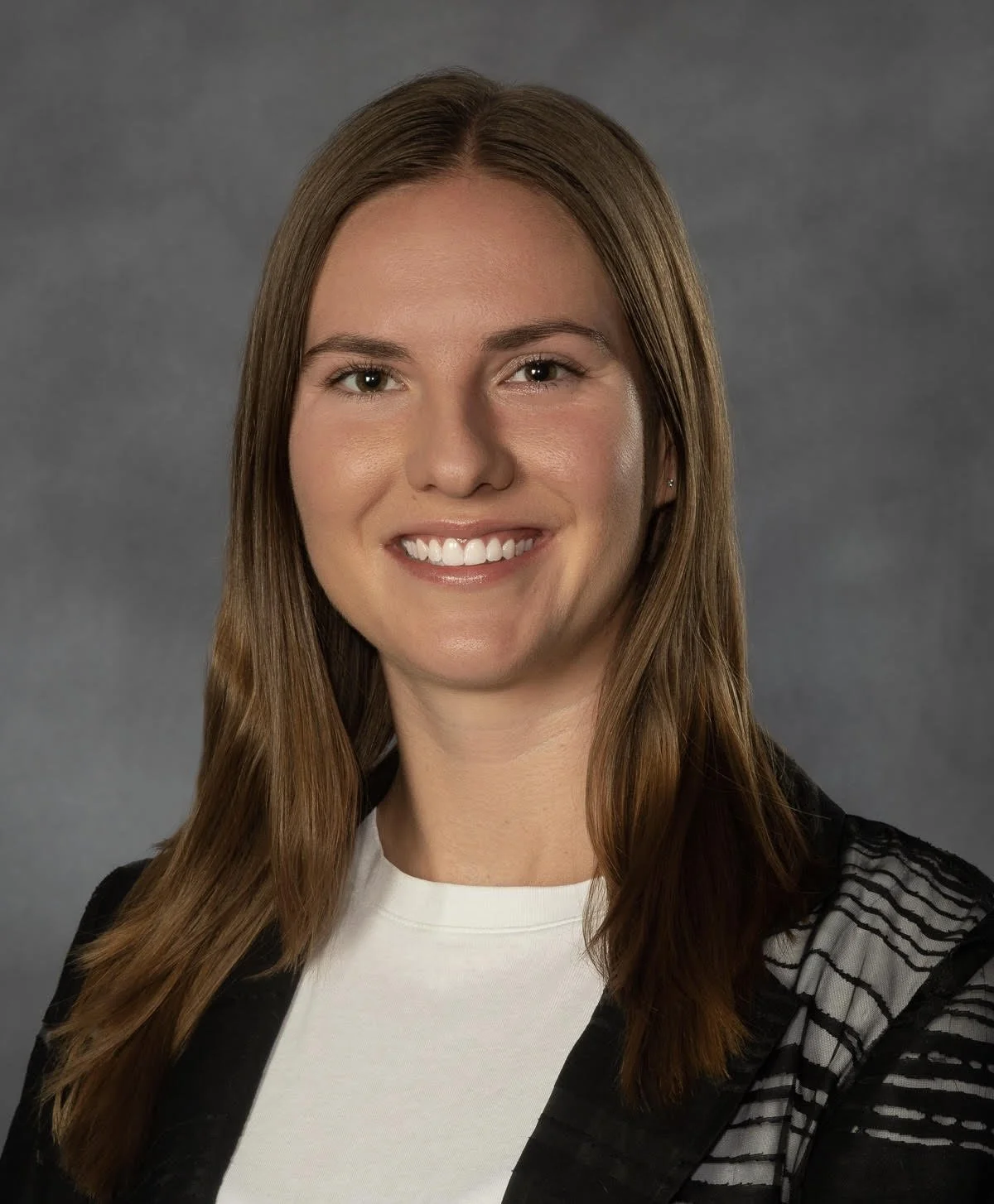 Professional headshot of a woman with shoulder-length brown hair, smiling, wearing a white top and a black blazer with striped detailing on the shoulder, against a gray background.