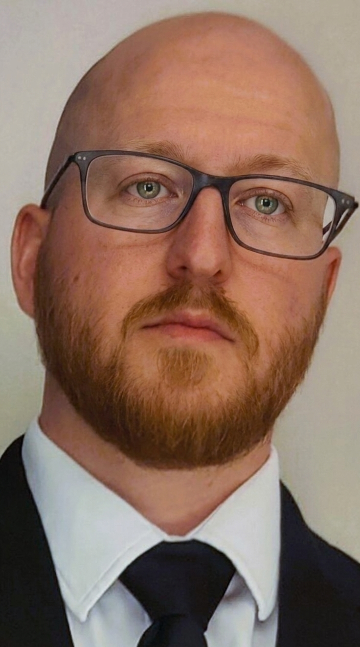 A man with a bald head, glasses, and a reddish-brown beard, dressed in a formal black suit, white shirt, and black tie.