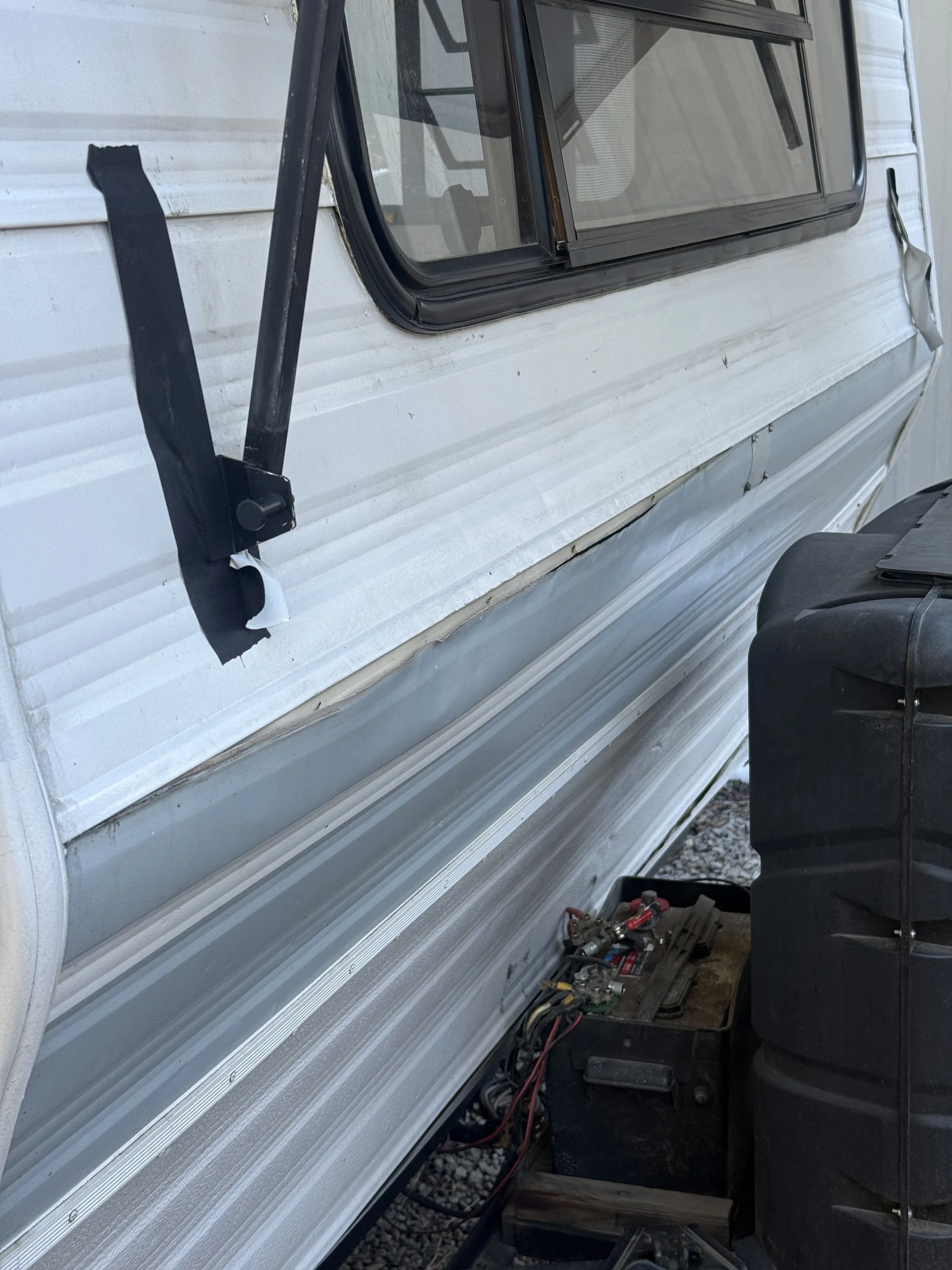 Close-up of a damaged camper trailer's exterior wall with a broken window and a black toolbox nearby.
