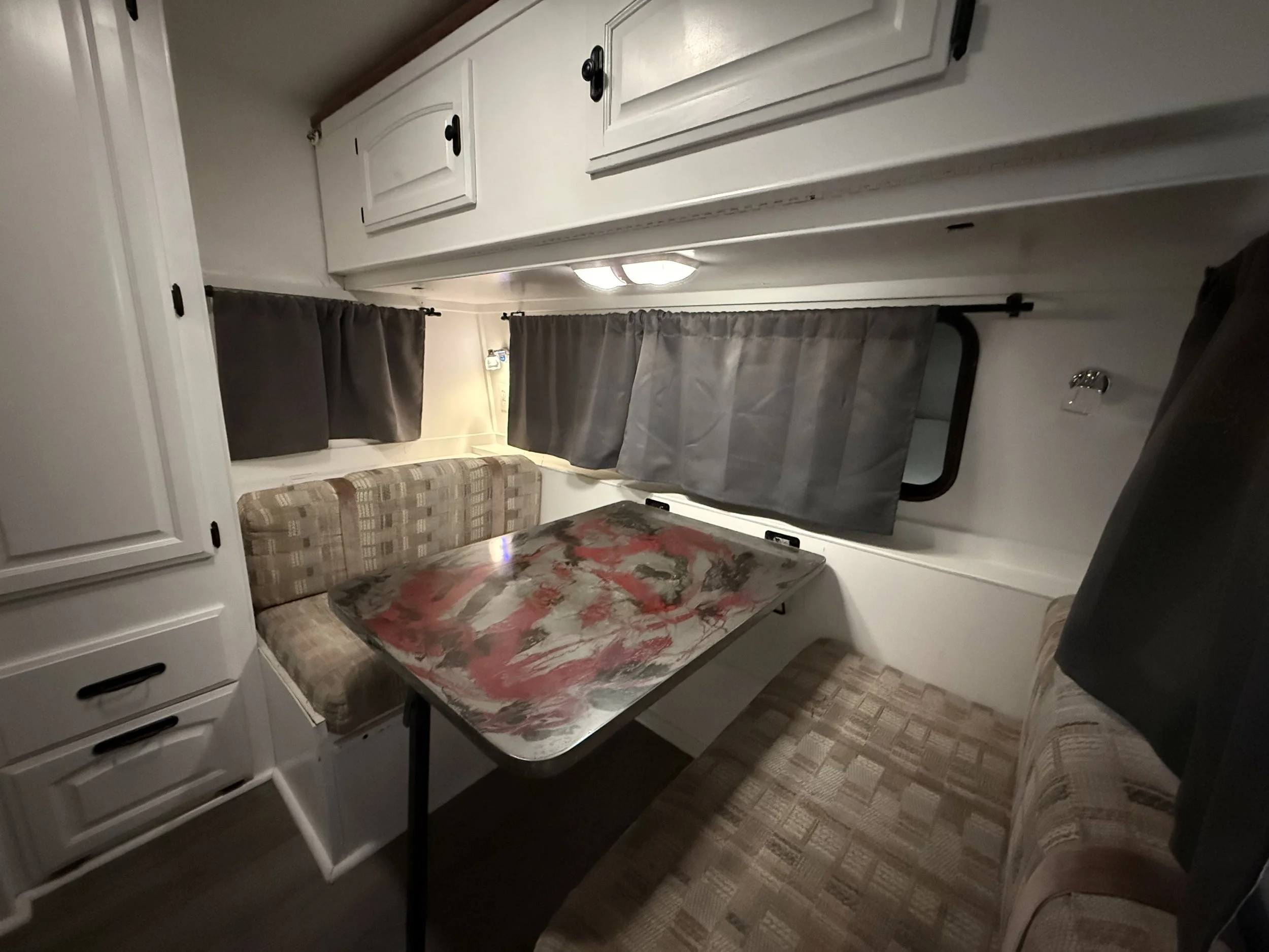 Interior of a small camper or RV after water damage repair with a dining area featuring a patterned upholstered bench on both sides of a table with a colorful abstract design, gray curtains over small windows, white cabinets overhead