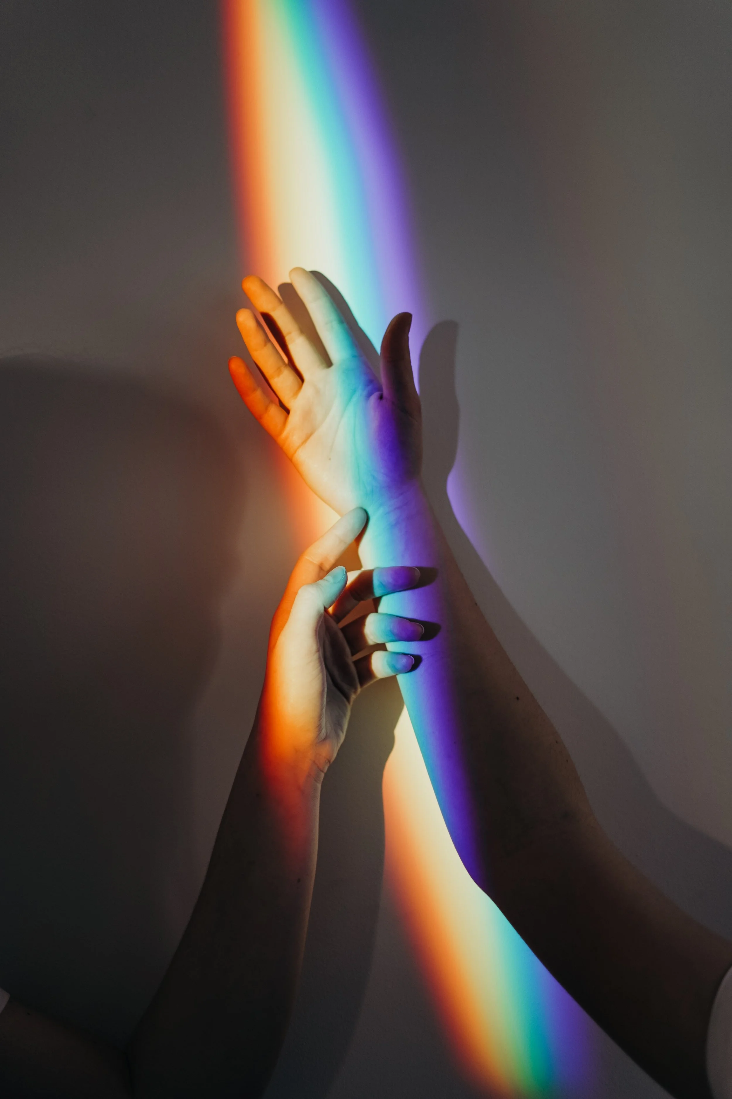 Two hands reaching towards each other with a rainbow light projected on them, creating colorful shadows on the wall.