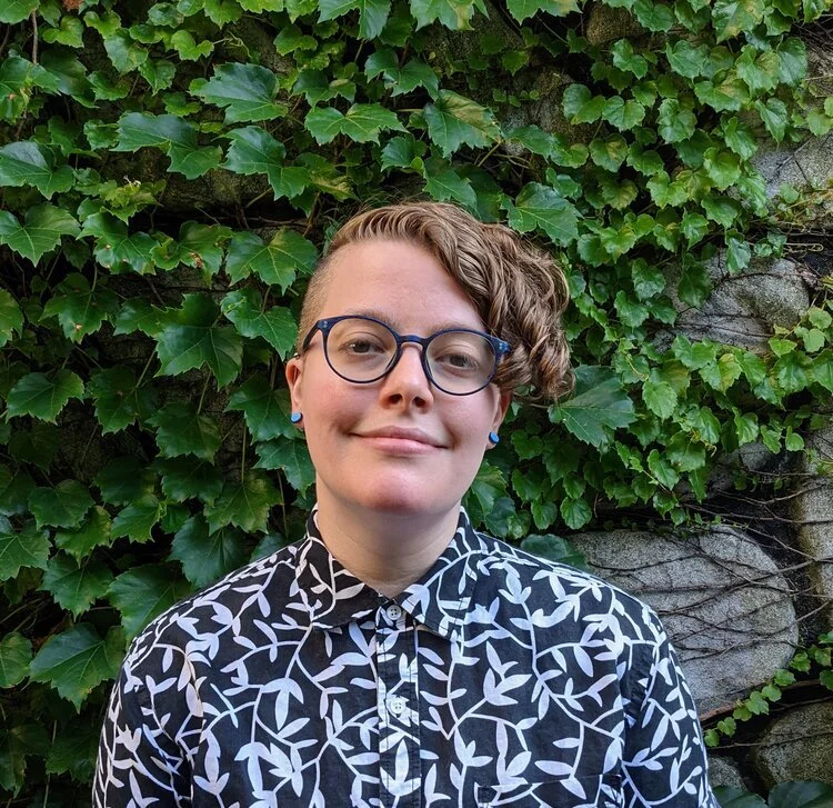 Person with short, curly hair, glasses, and earrings, standing in front of green ivy on a stone wall, wearing a black and white patterned shirt, smiling softly.