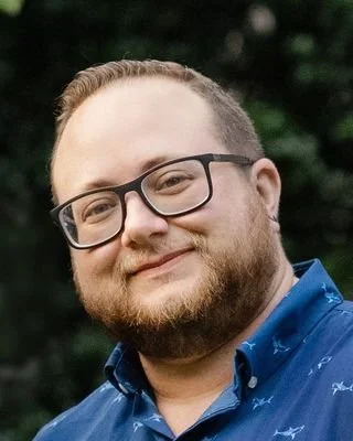 A man with glasses and a beard smiling outdoors, wearing a blue shirt.