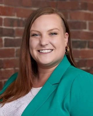 Portrait of a young woman with long red hair, smiling, wearing a teal blazer and white top, standing in front of a brick wall.