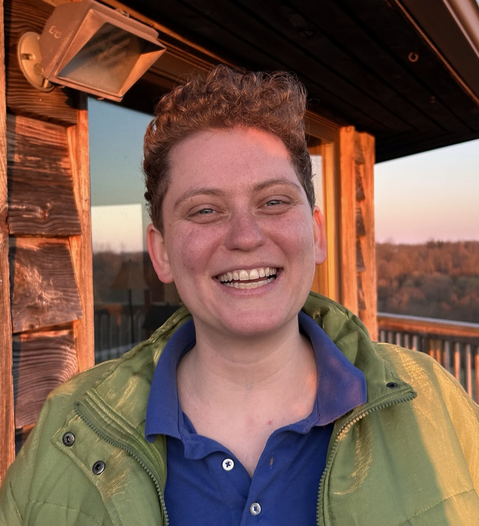 Smiling person with short curly hair, wearing a green jacket and blue polo shirt, standing on a wooden deck with a sunset view in the background.