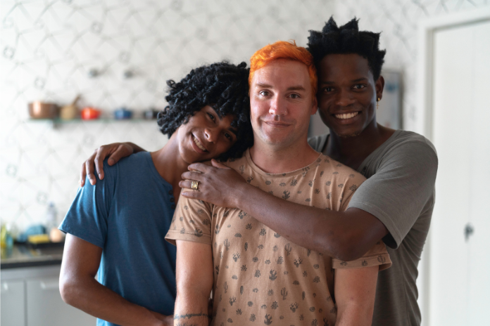Three people smiling and hugging in a modern kitchen with patterned wallpaper.