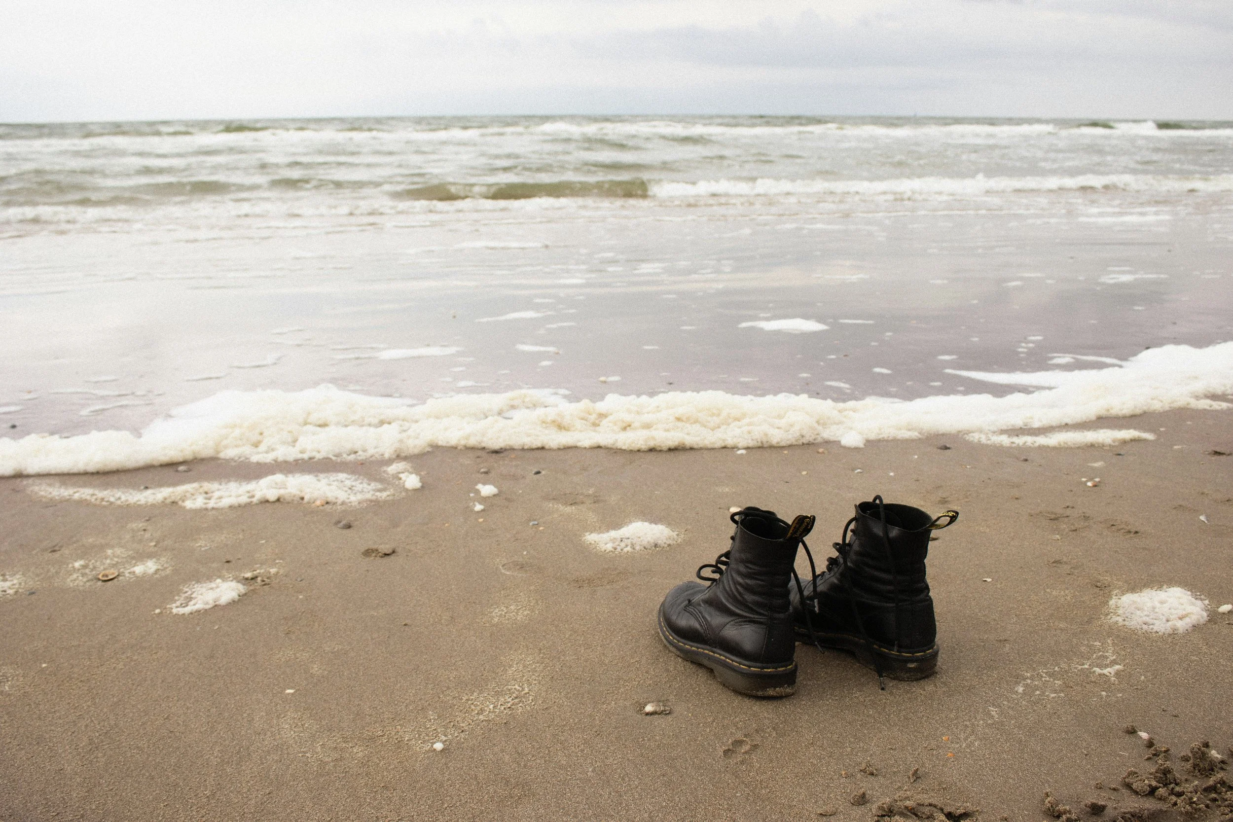 A pair of black boots on the sandy beach near the shoreline with ocean waves in the background.