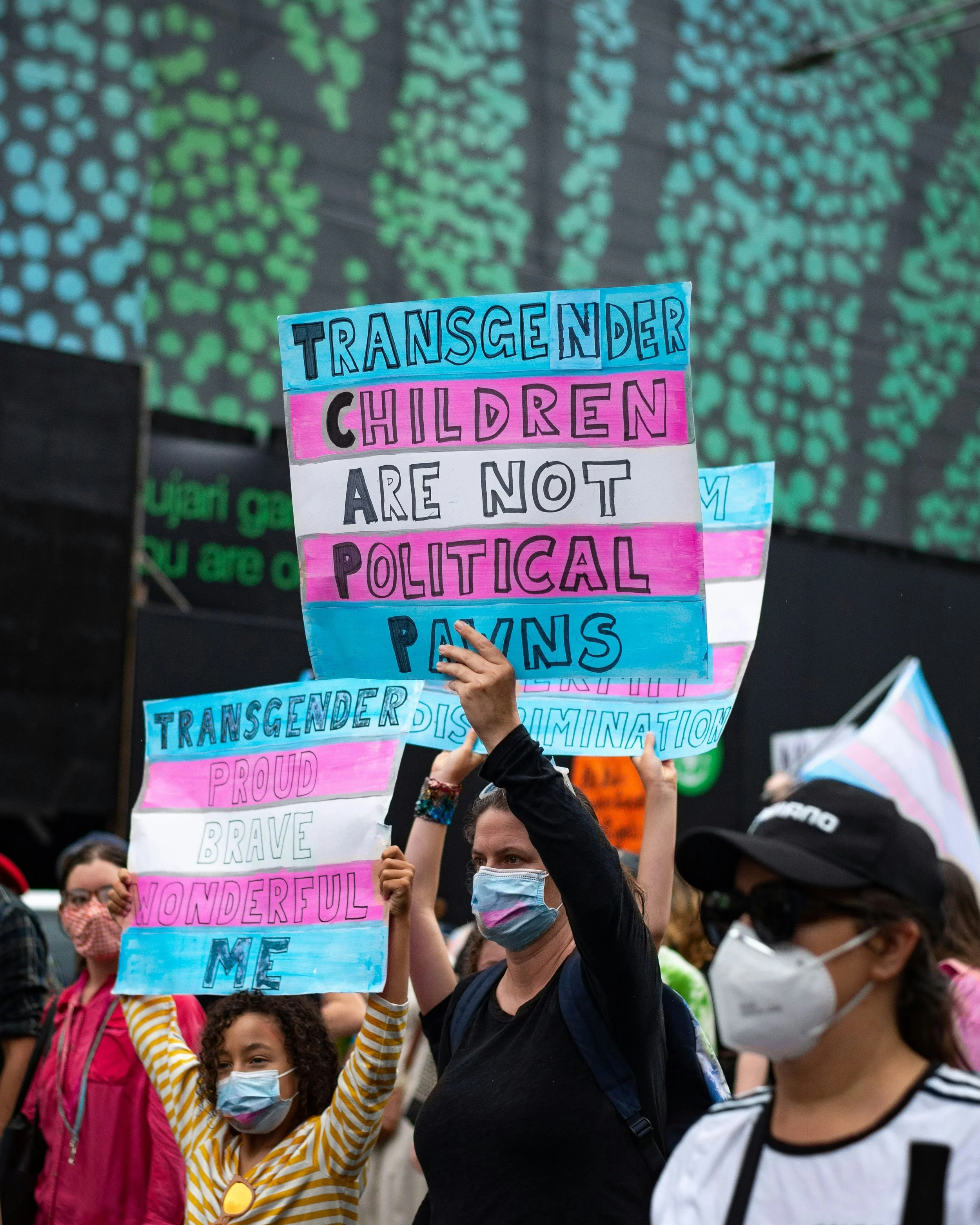Group of people, some wearing masks, holding signs supporting transgender children at a rally or march. The signs display messages like "Transgender children are not political pawns" and "Transgender proud, brave, wonderful me."