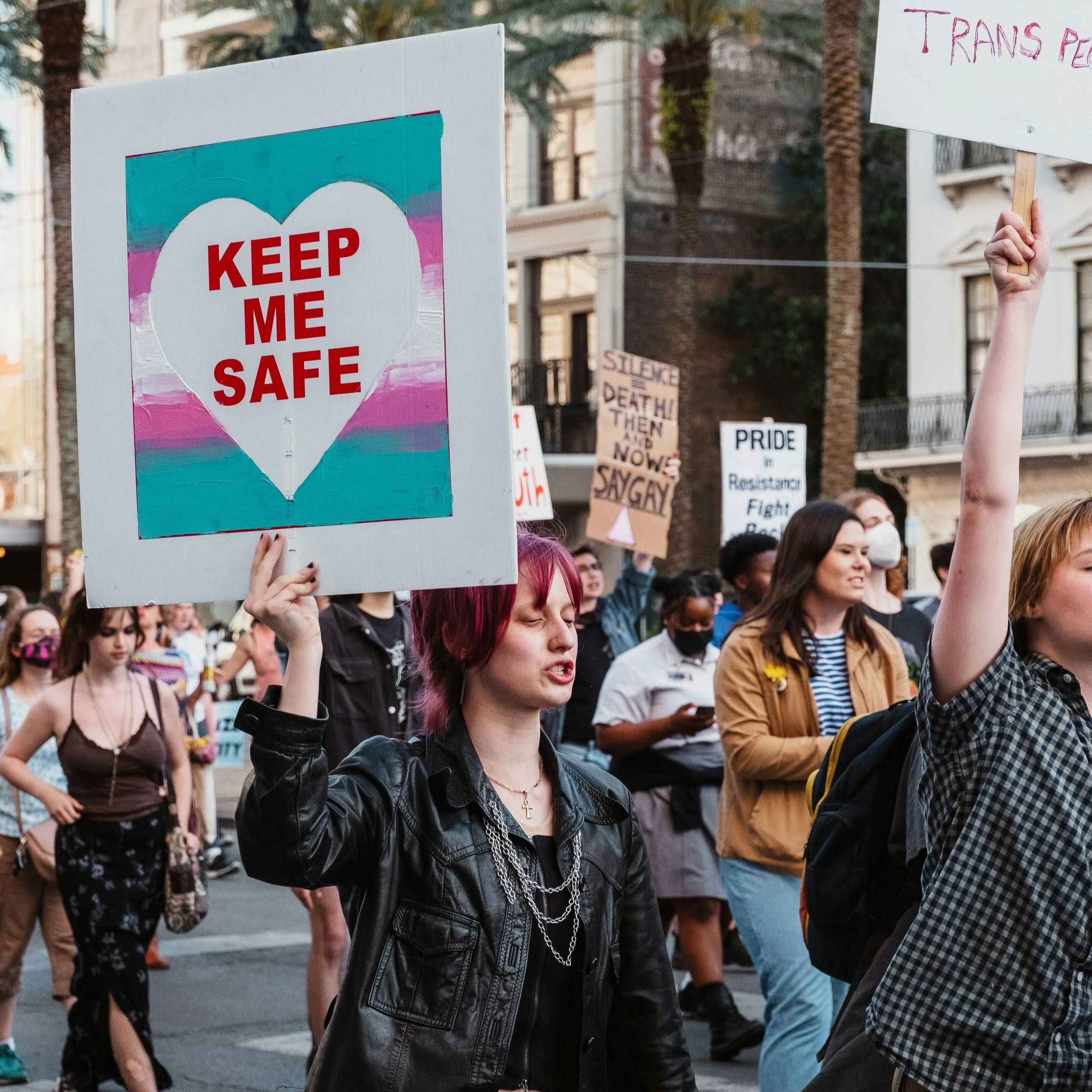 A woman with short pink hair holding a sign with a white heart and the words 'KEEP ME SAFE' in red during a protest or rally.