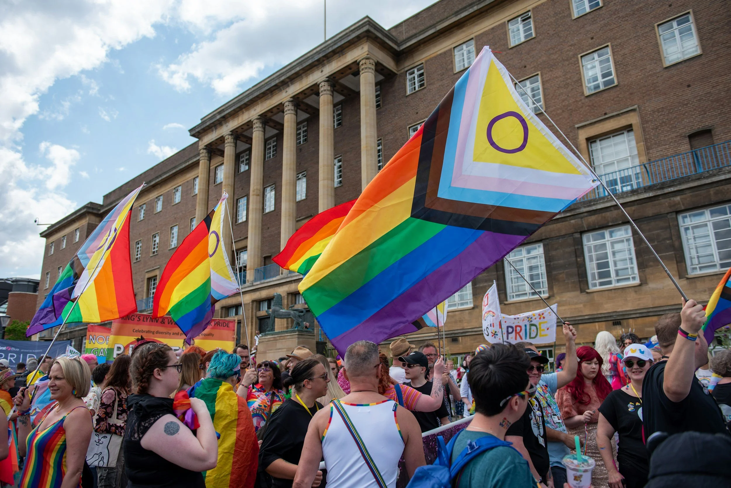 LGBTQ+ pride parade with multiple rainbow flags and a crowd of people celebrating outdoors in front of a large brick building.