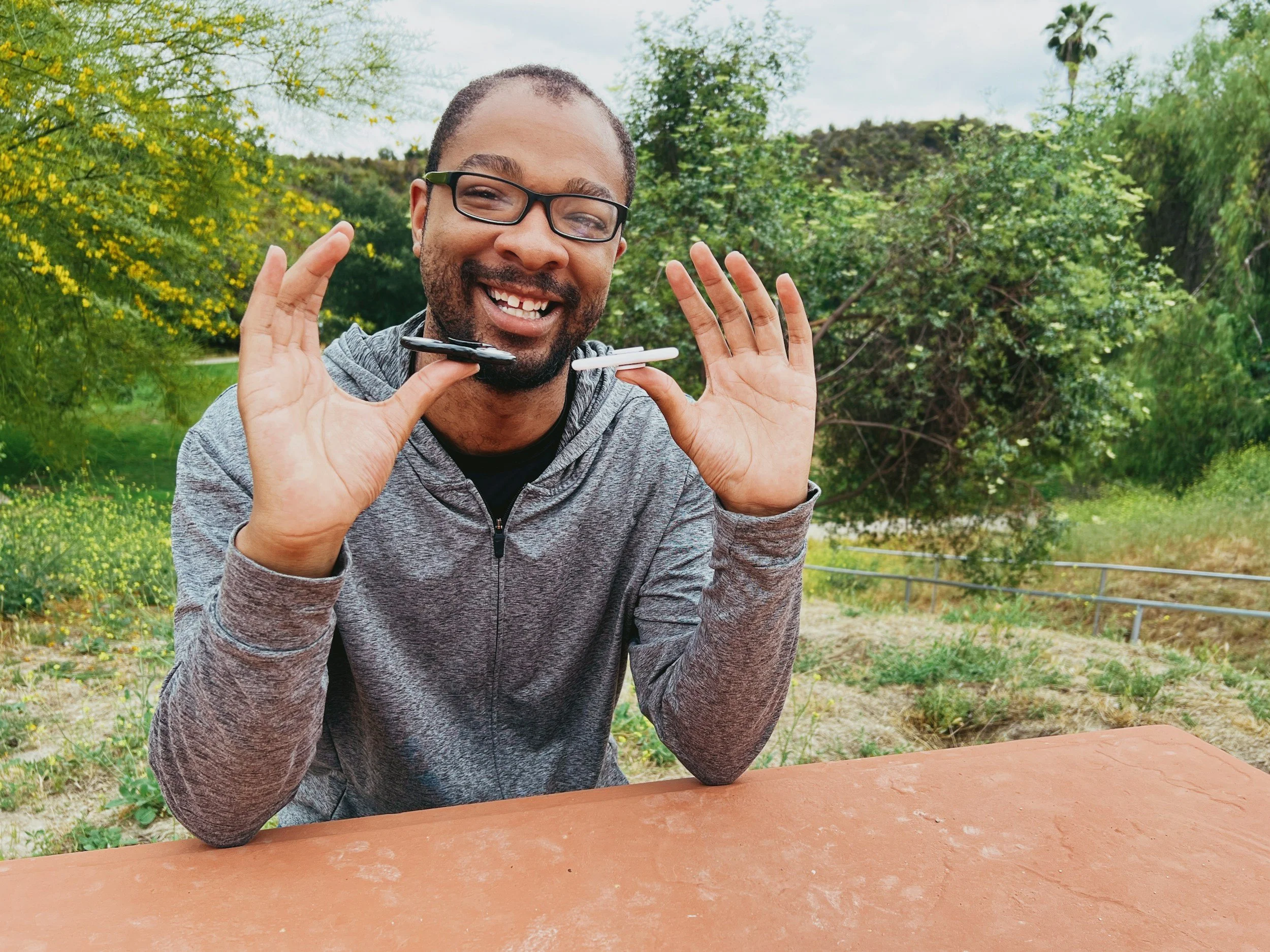 Man with glasses smiling outdoors, holding a pen horizontally at chin level, with lush green trees in the background.