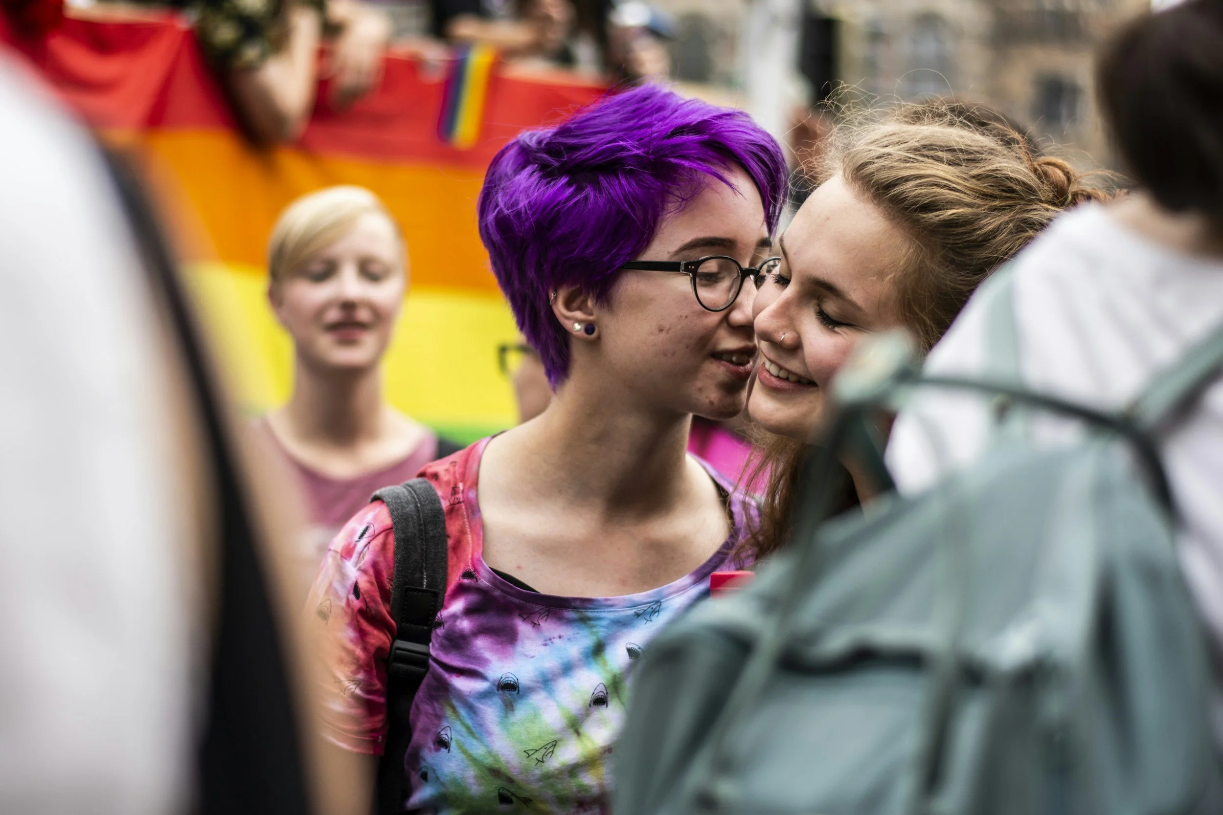 Two women, one with purple hair and glasses and the other with brown hair, are close together and smiling at a pride event with a rainbow flag in the background.
