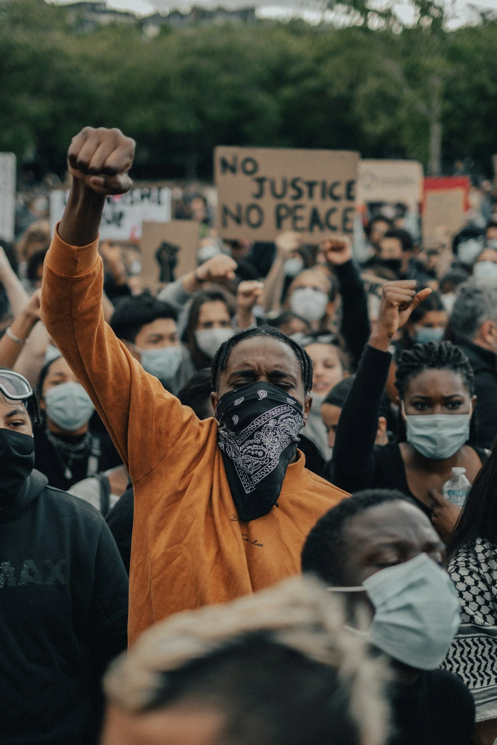 A diverse crowd of protesters, many wearing face masks, gathered outdoors holding signs, with one sign reading 'NO JUSTICE NO PEACE.' A person in the foreground is raising a clenched fist, symbolizing protest or defiance.