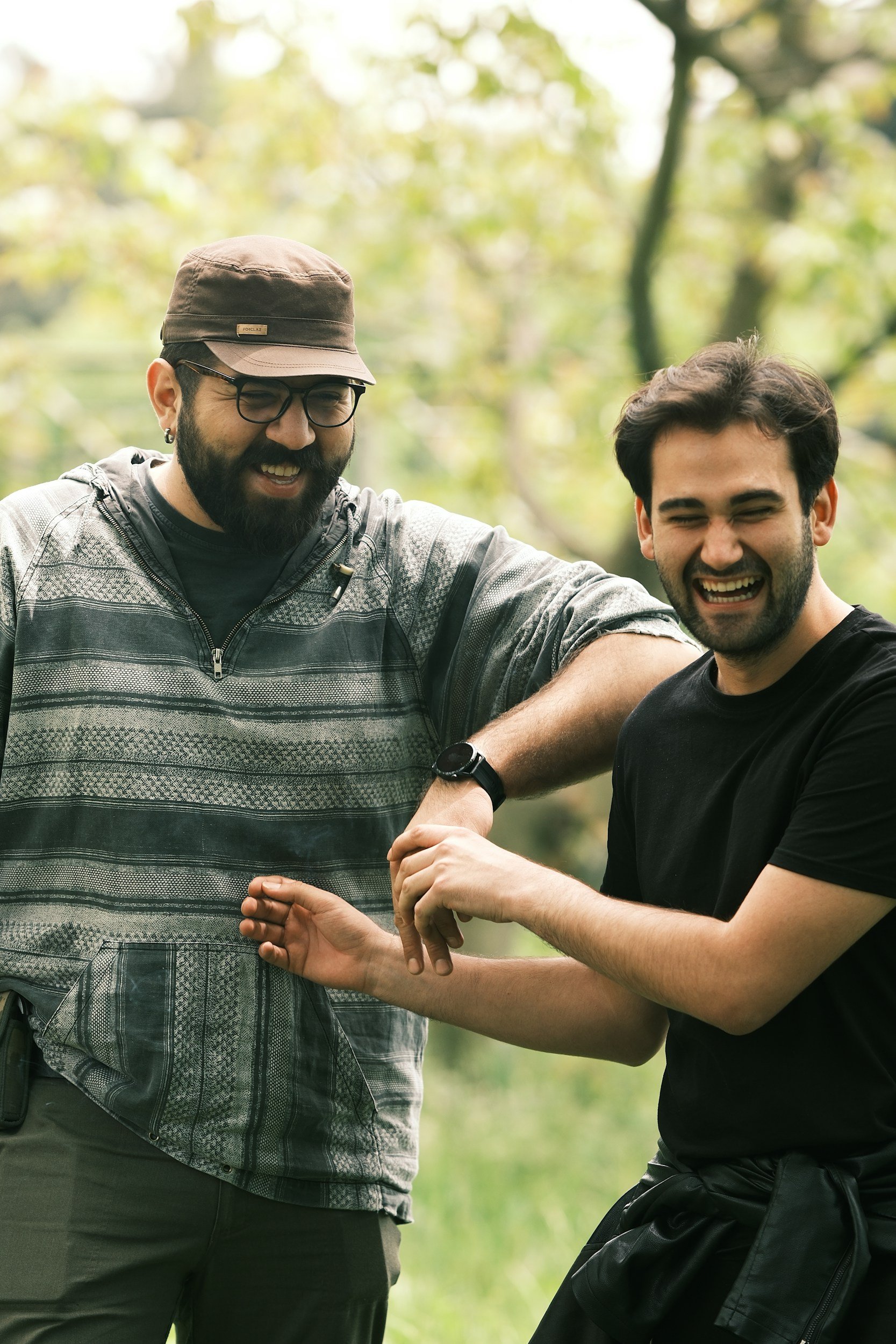 Two men are smiling and laughing outdoors in a green forest, with one man's arm around the other's shoulder.