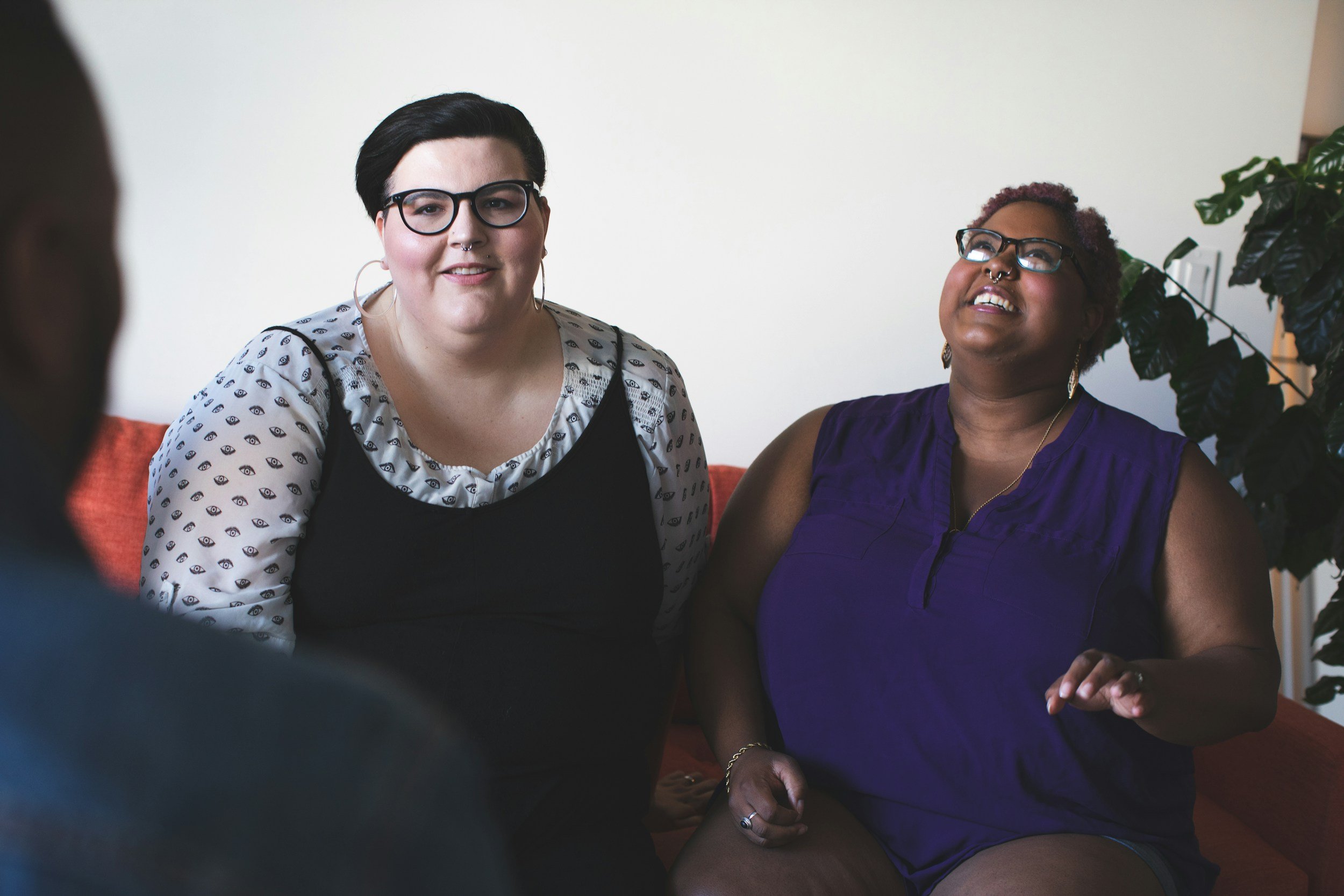 Two women sitting on a red couch, engaging in a conversation. One woman is wearing glasses and a black dress over a patterned shirt, while the other is wearing glasses and a sleeveless purple top.