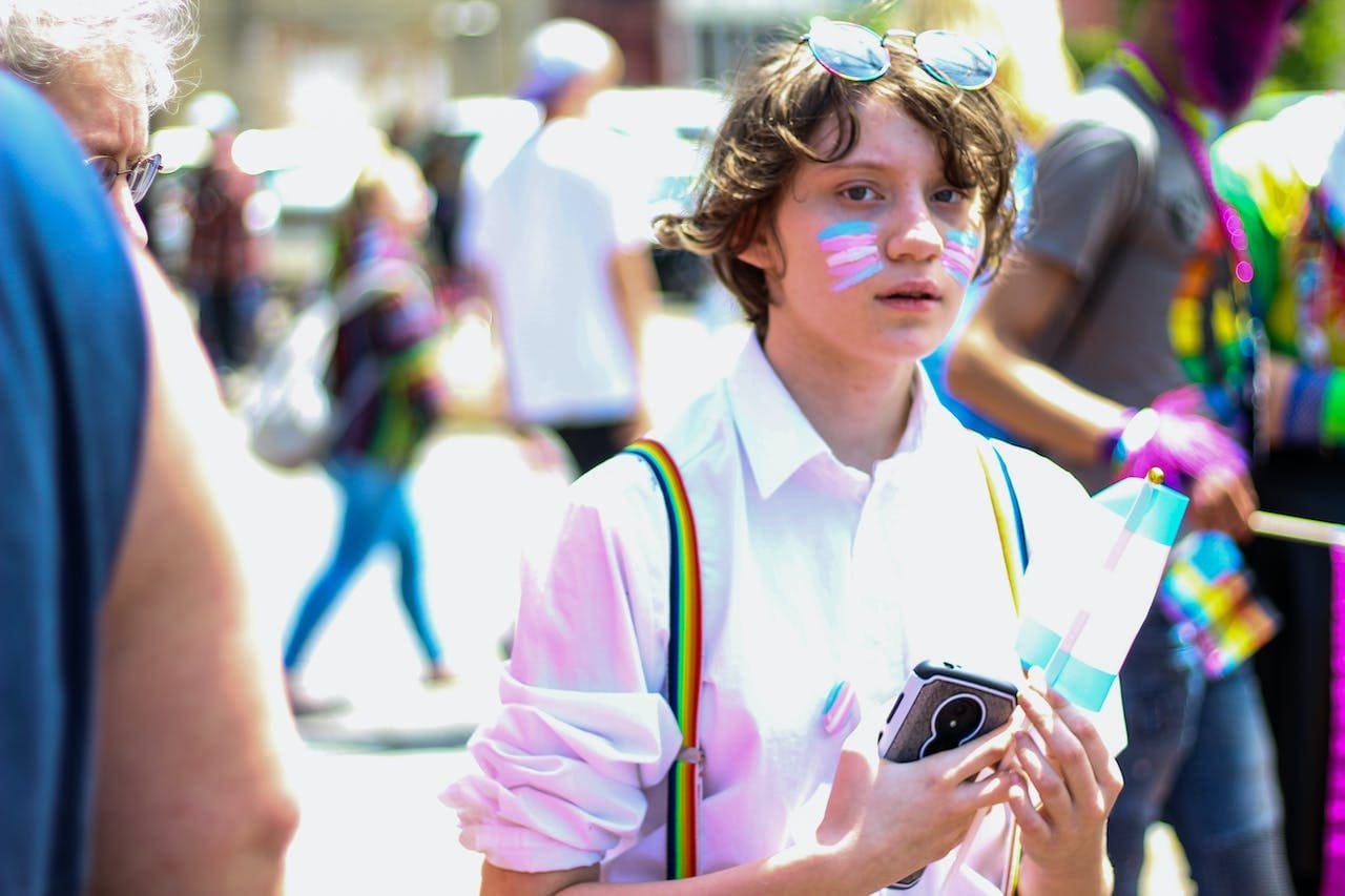 Young person with rainbow face paint, rainbow suspenders, and sunglasses on head, holding a phone, paper, and a small flag at a pride parade.