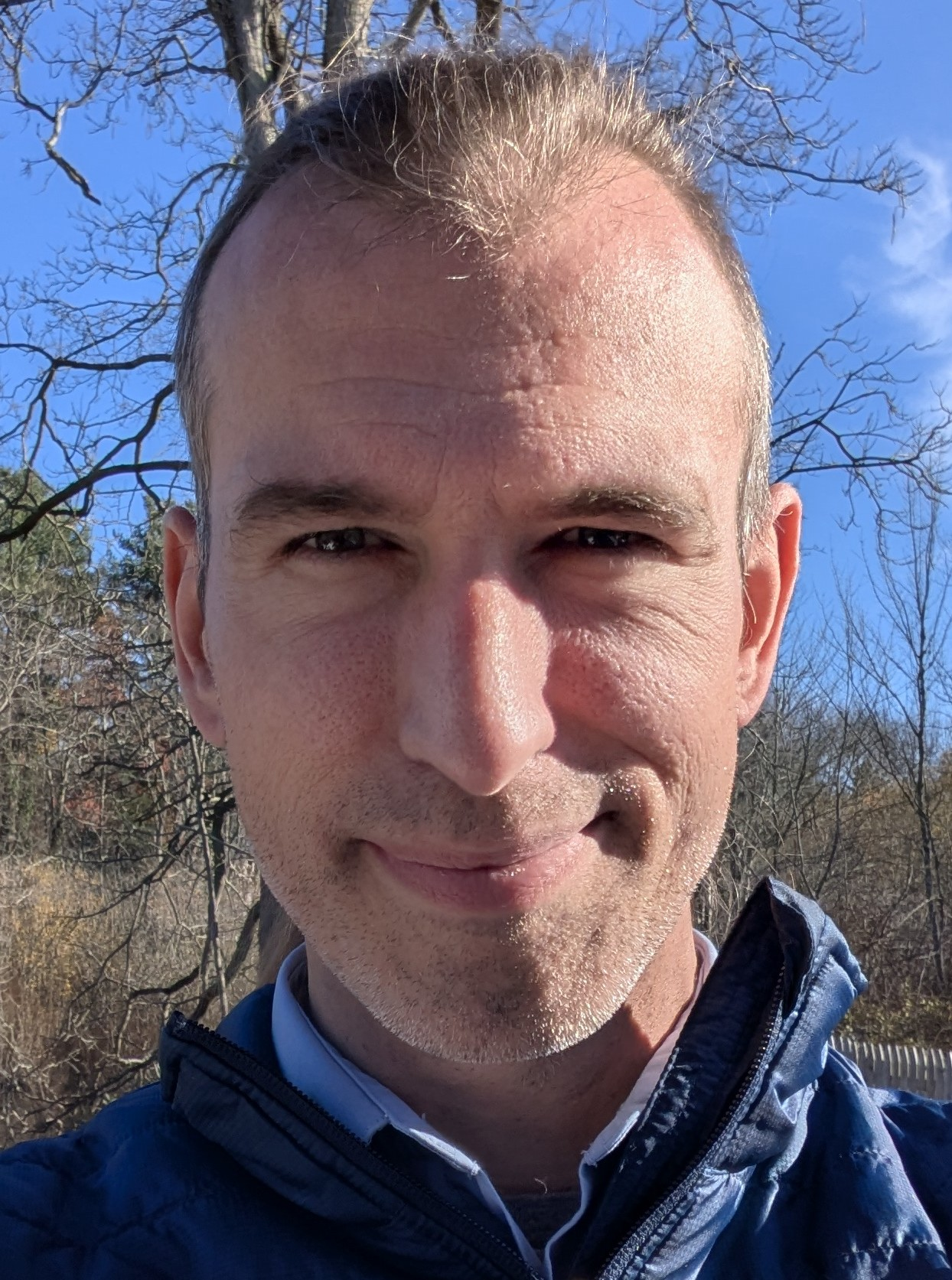 A close-up of a person outdoors, wearing a blue jacket, with bare trees and a blue sky in the background.