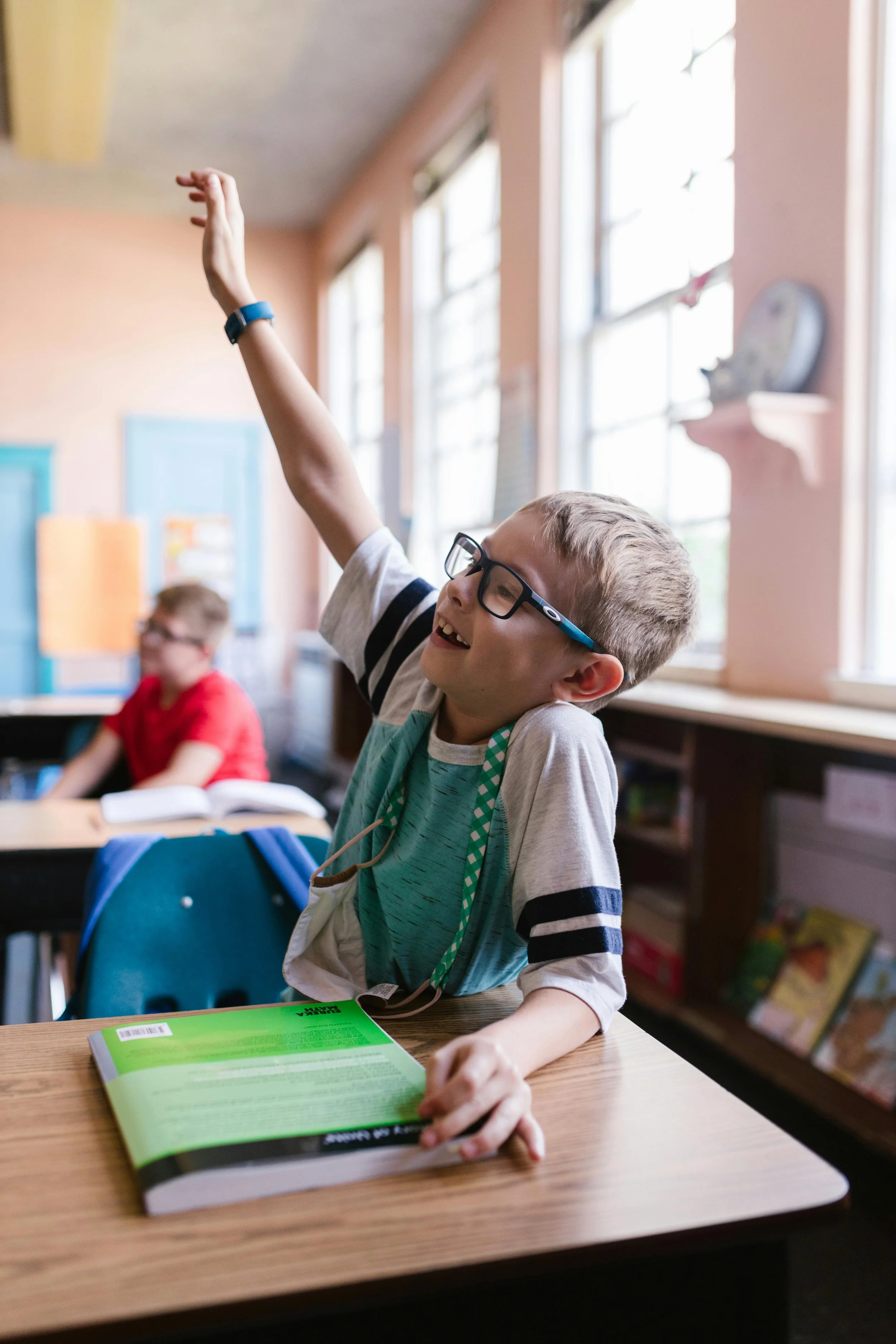 A happy young boy with glasses raising his hand in a classroom, with another child in the background, sitting at desks and books on the tables.