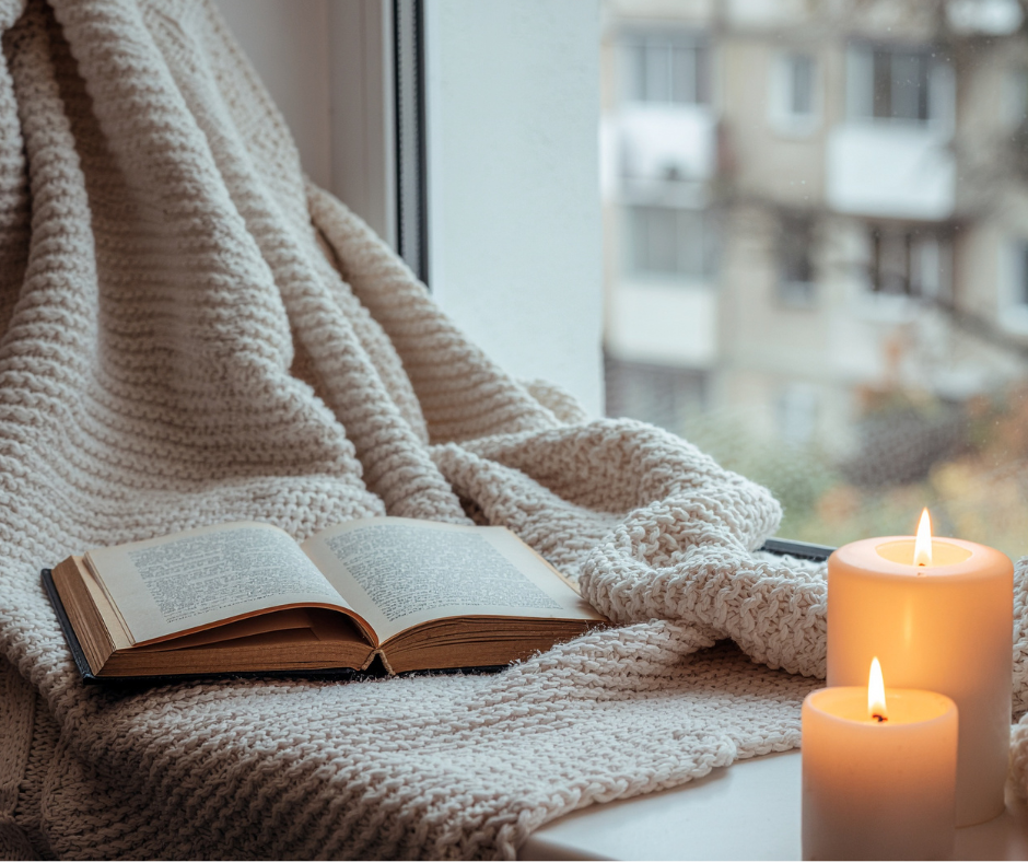 An open book resting on a cozy, knitted blanket by a window with lit candles nearby. Bright Earth Wellness Co candle creating a calm, wellness-focused home environment