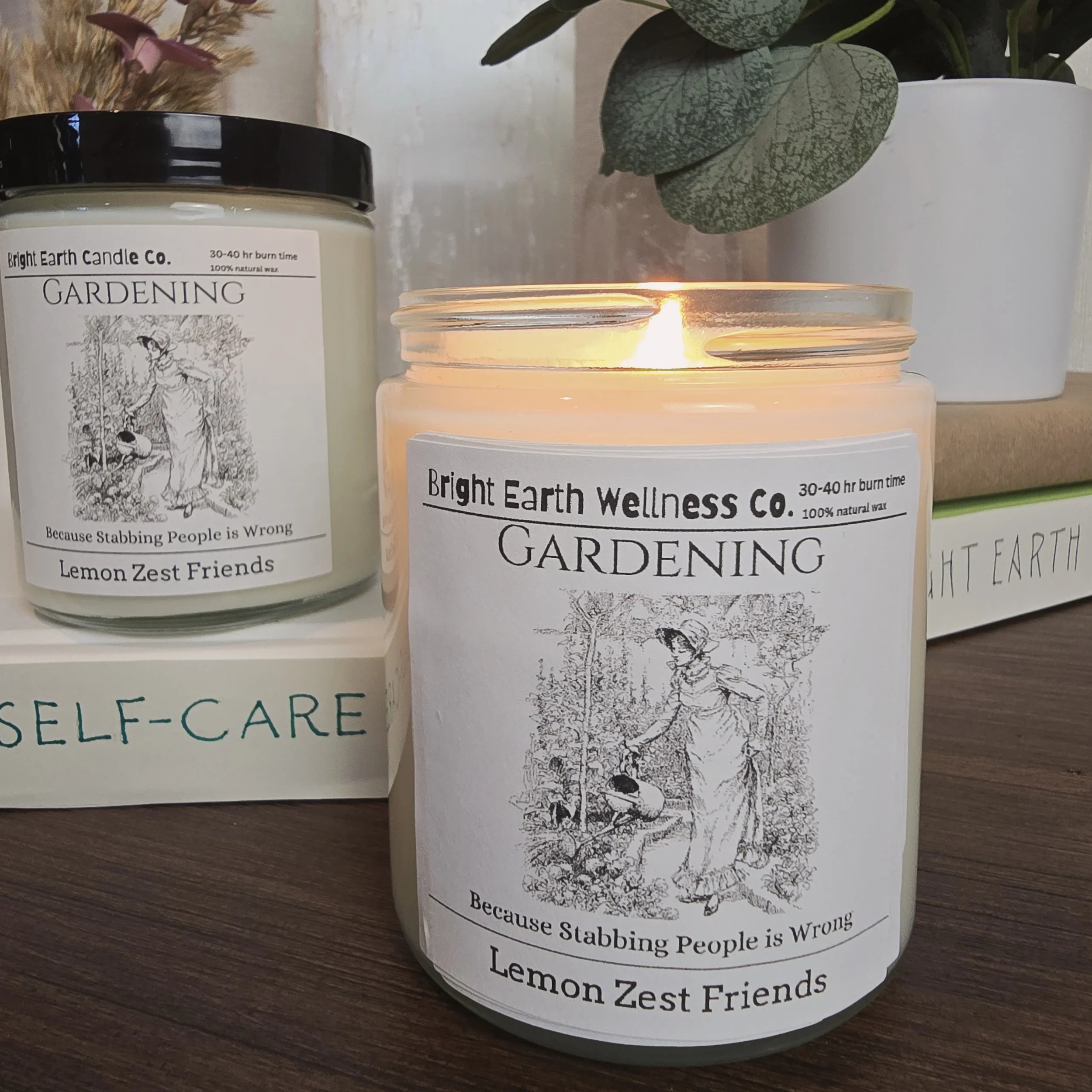 Two candles from Bright Earth Wellness Co. labeled 'GARDENING'. One candle is lit and emits a warm glow. The labels feature a black-and-white illustration of a woman gardening, with a tagline 'Because Stabbing People is Wrong' and the scent named 'Lemon Zest Friends'. A potted plant is also visible in the background.