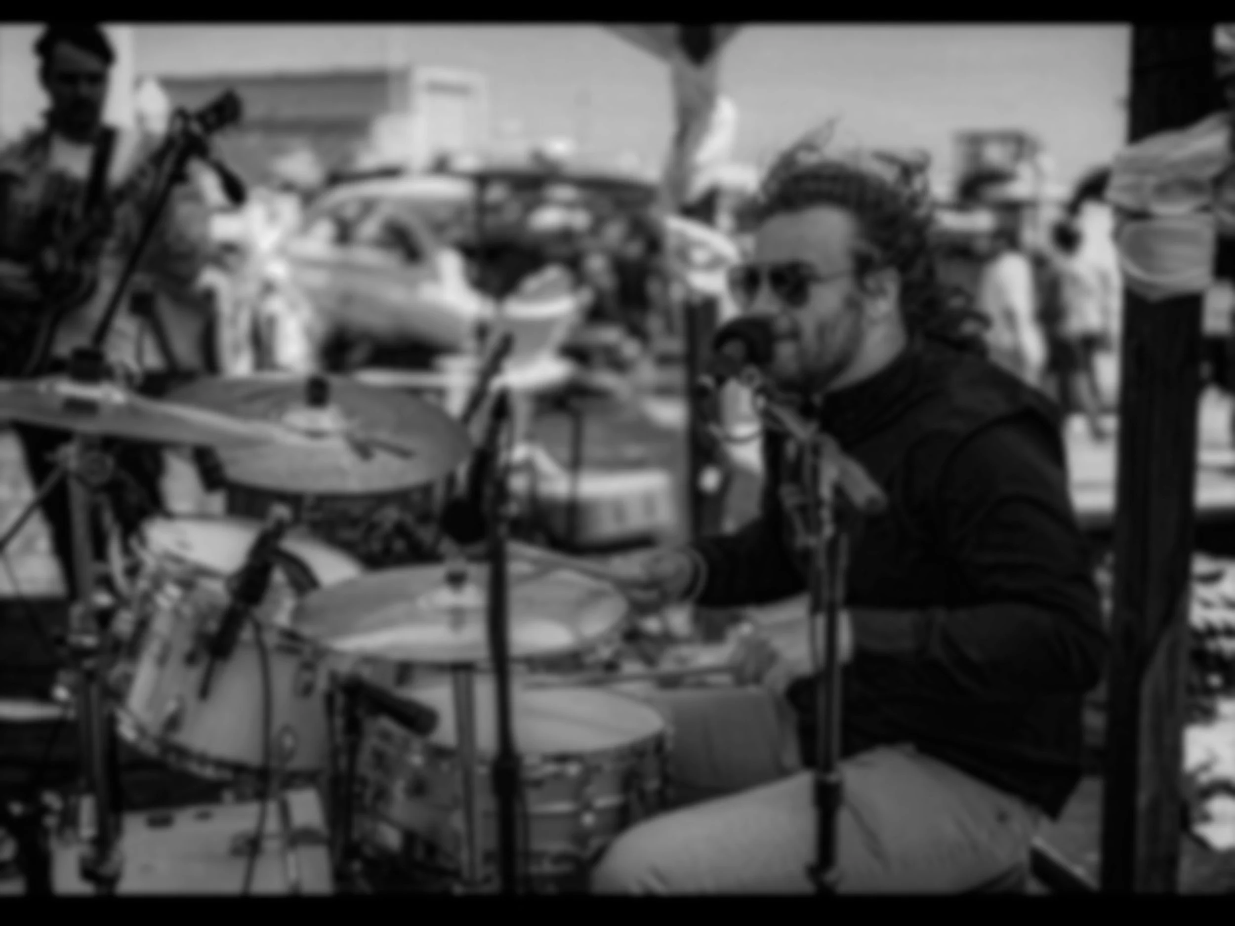 A man playing drums outdoors at a busy market or street event, with cars and people in the background.