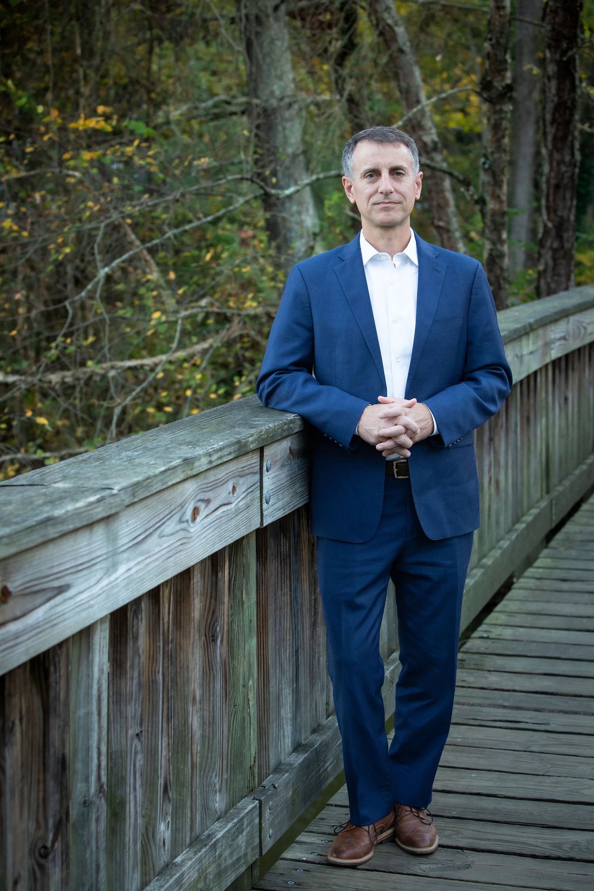 A man in a blue suit and white shirt standing on a wooden bridge outdoors with trees and foliage in the background.