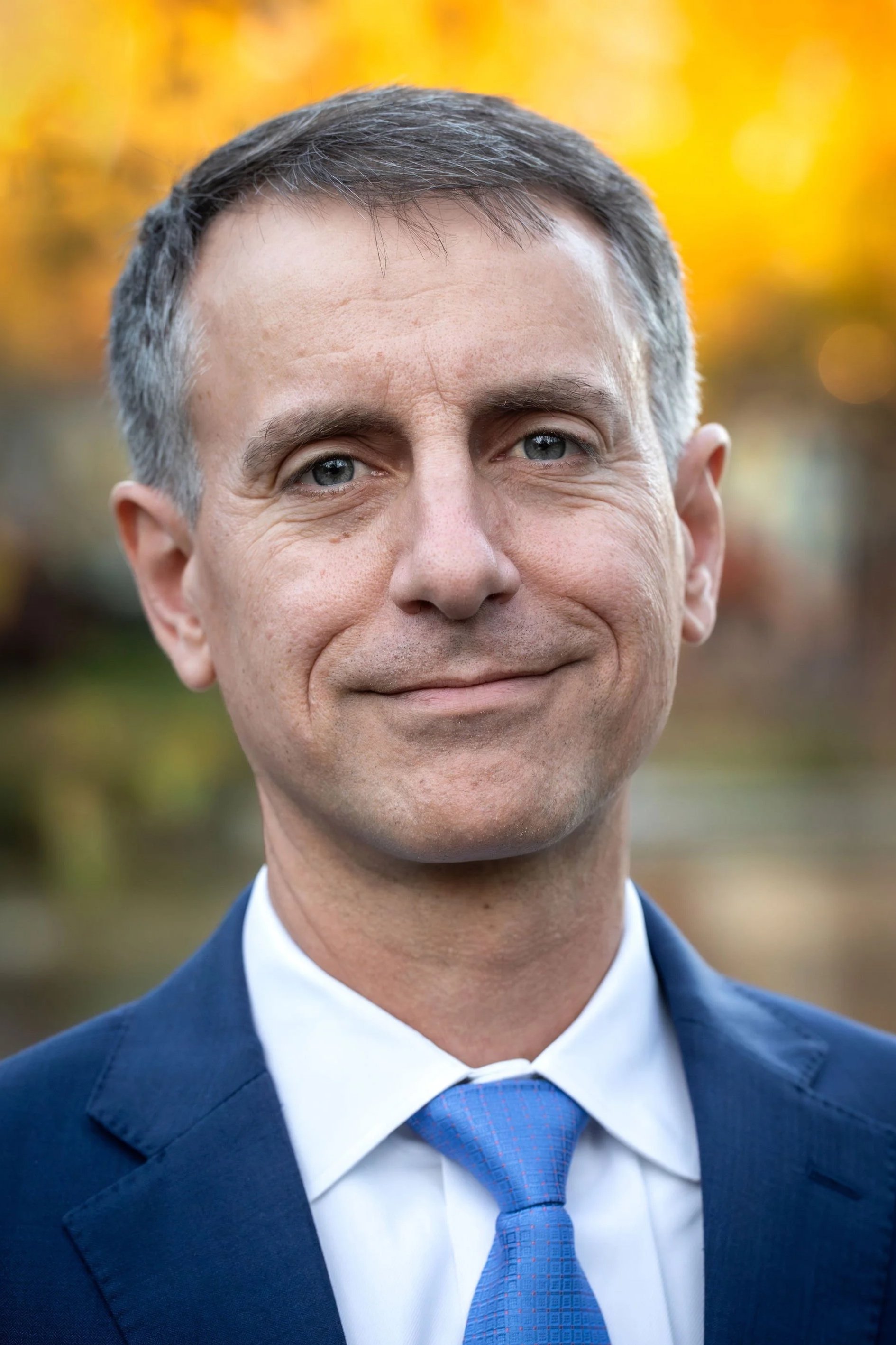 Close-up of a middle-aged man in a blue suit and tie, smiling outdoors with blurred autumn foliage in the background.