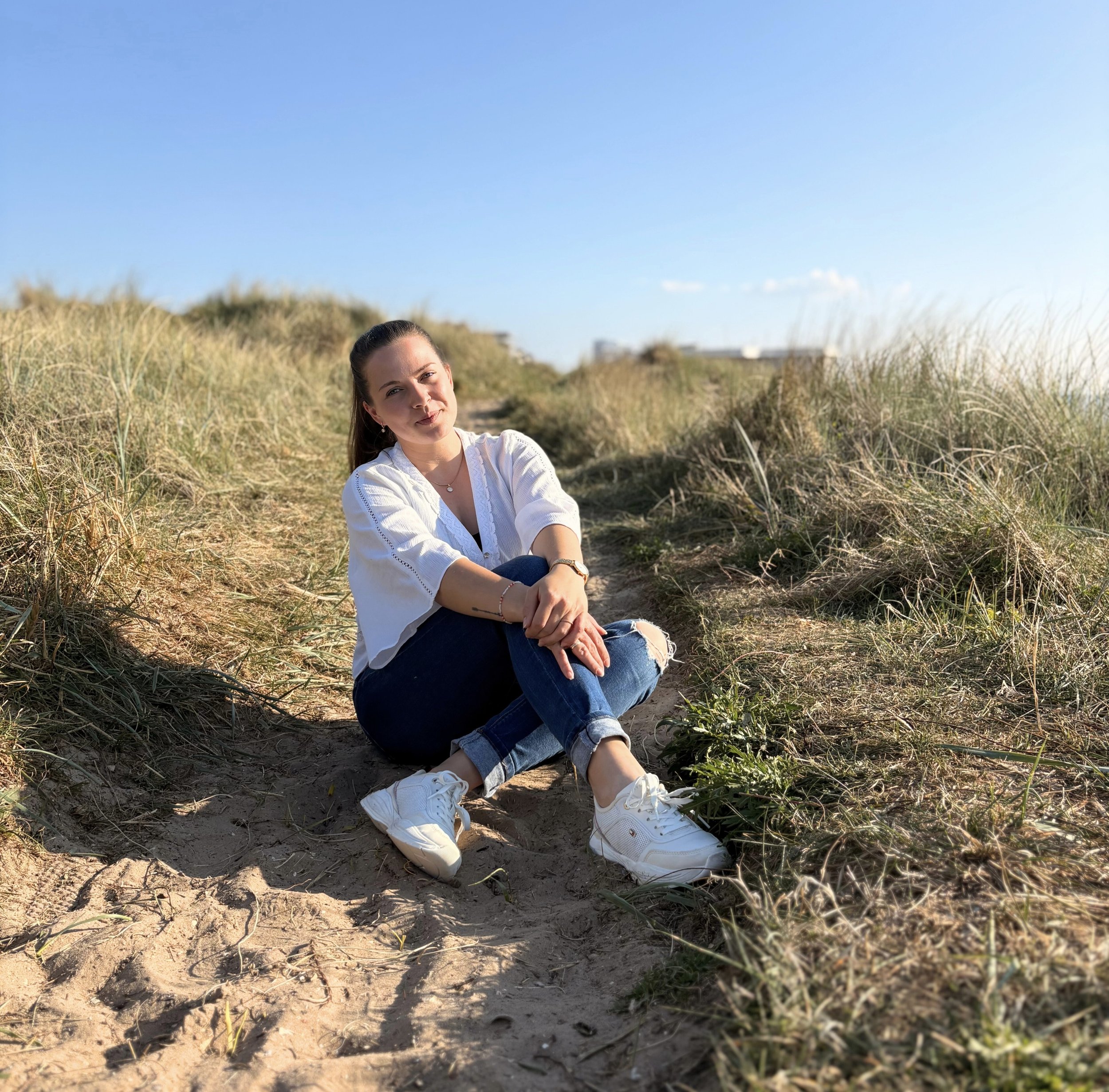 weiße Frau sitzt lächelnd in Dünen. Sie hat einen hohen Zopf, trägt weiße Schuhe, eine dunkle Jeans und ein weißes Shirt. Sie