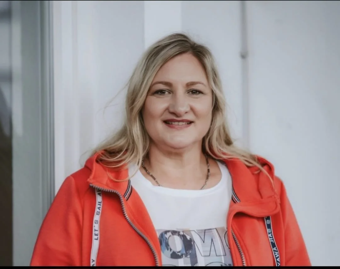 Frau mit langen blonden Haaren schaut frontal lächelnd in die Kamera. Sie trägt ein weißes Tshirt mit Print, sowie eine korallfarbene Jacke.