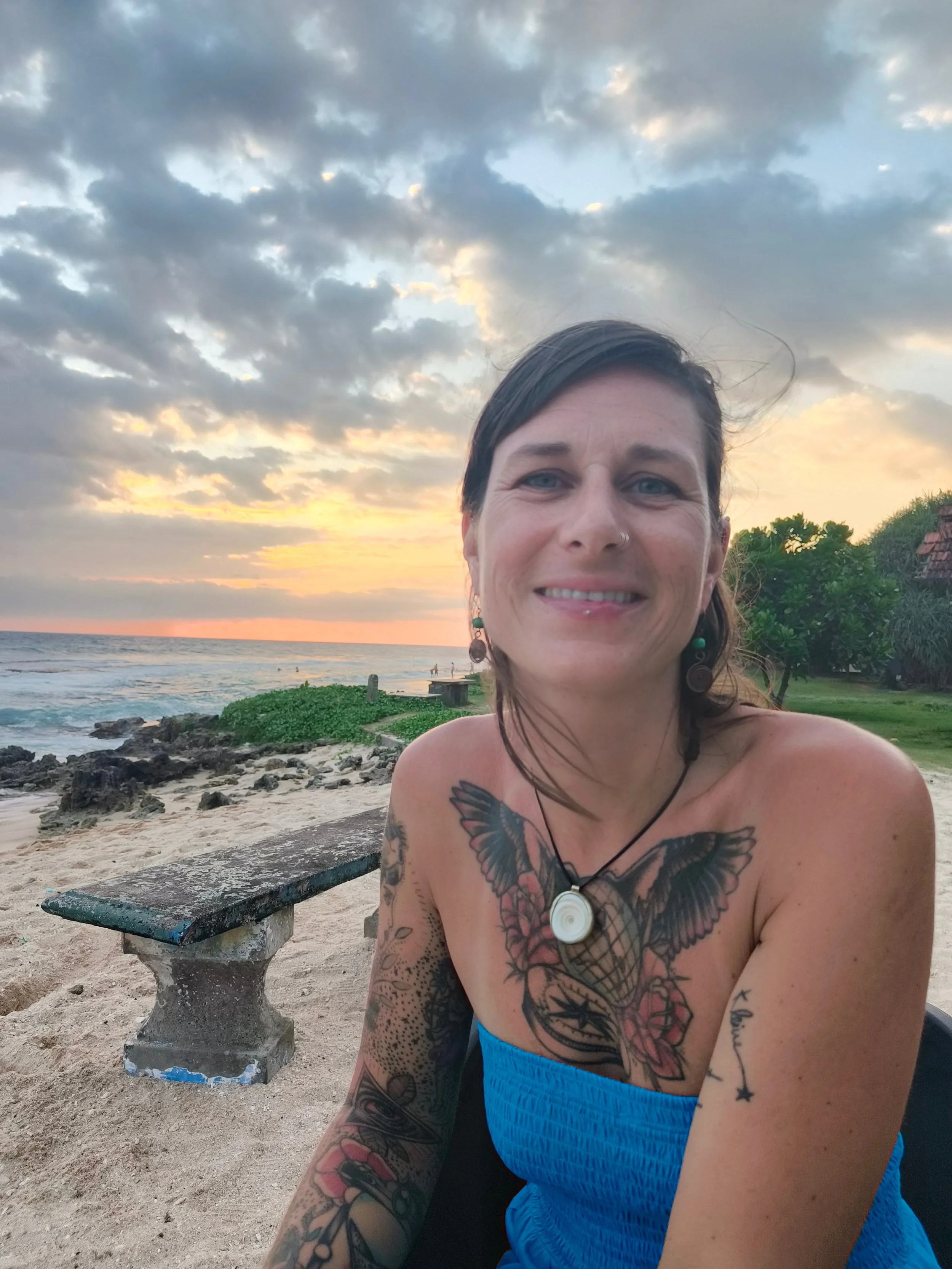Tättowierte Frau mit braunen Haaren und Seitenscheitel sitzt am Strand und trägt ein blaues Top mit einer markanten Kette um den Hals.