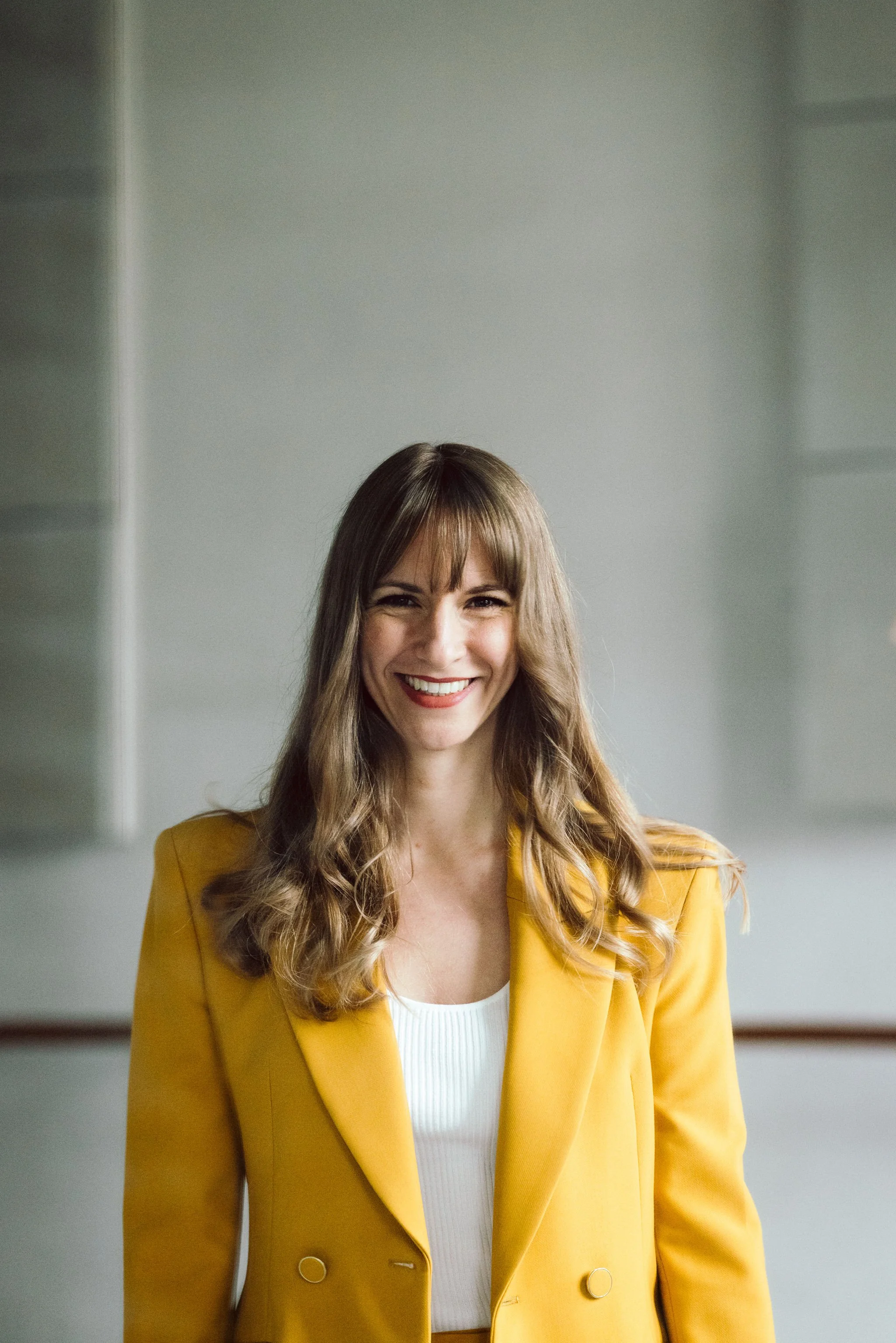 Junge Frau mit braunen langen Haaren und gelben Blazer schaut lachend in die Kamera