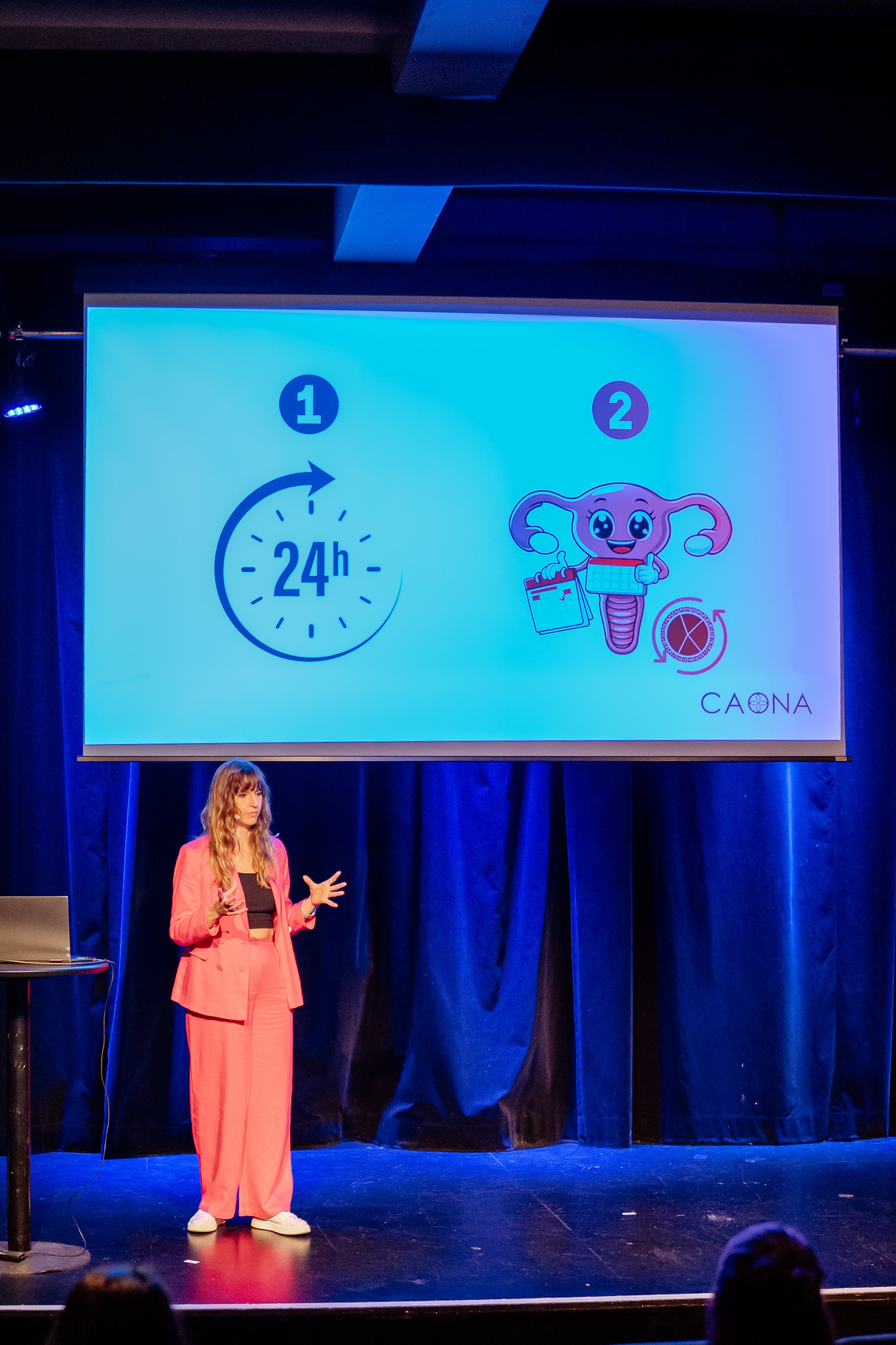 Frau in pinkem Anzug hält eine Rede auf der Bühne. Im Hintergrund Präsentation mit Zeit-Symbol und Grafik des weiblichen Uterus