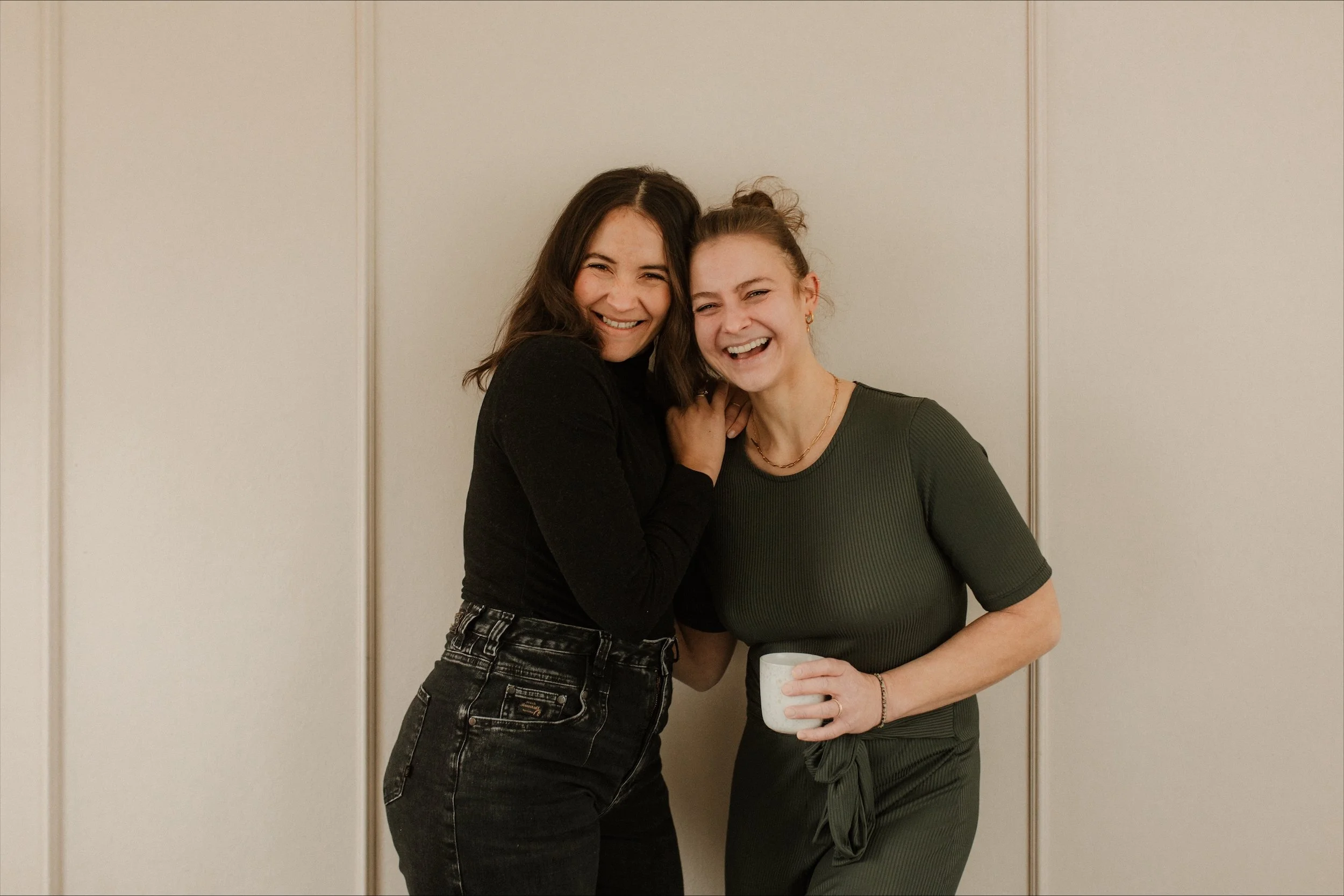 Zwei Frauen stehen nebeneinander und schauen lächelnd in die Kamera. Auf der linken Seite mit dunklen Haaren und schwarzem Shirt und auf der rechten Seite mit blonden Haaren im Dutt gebunden mit Kaffeetasse in der Hand.