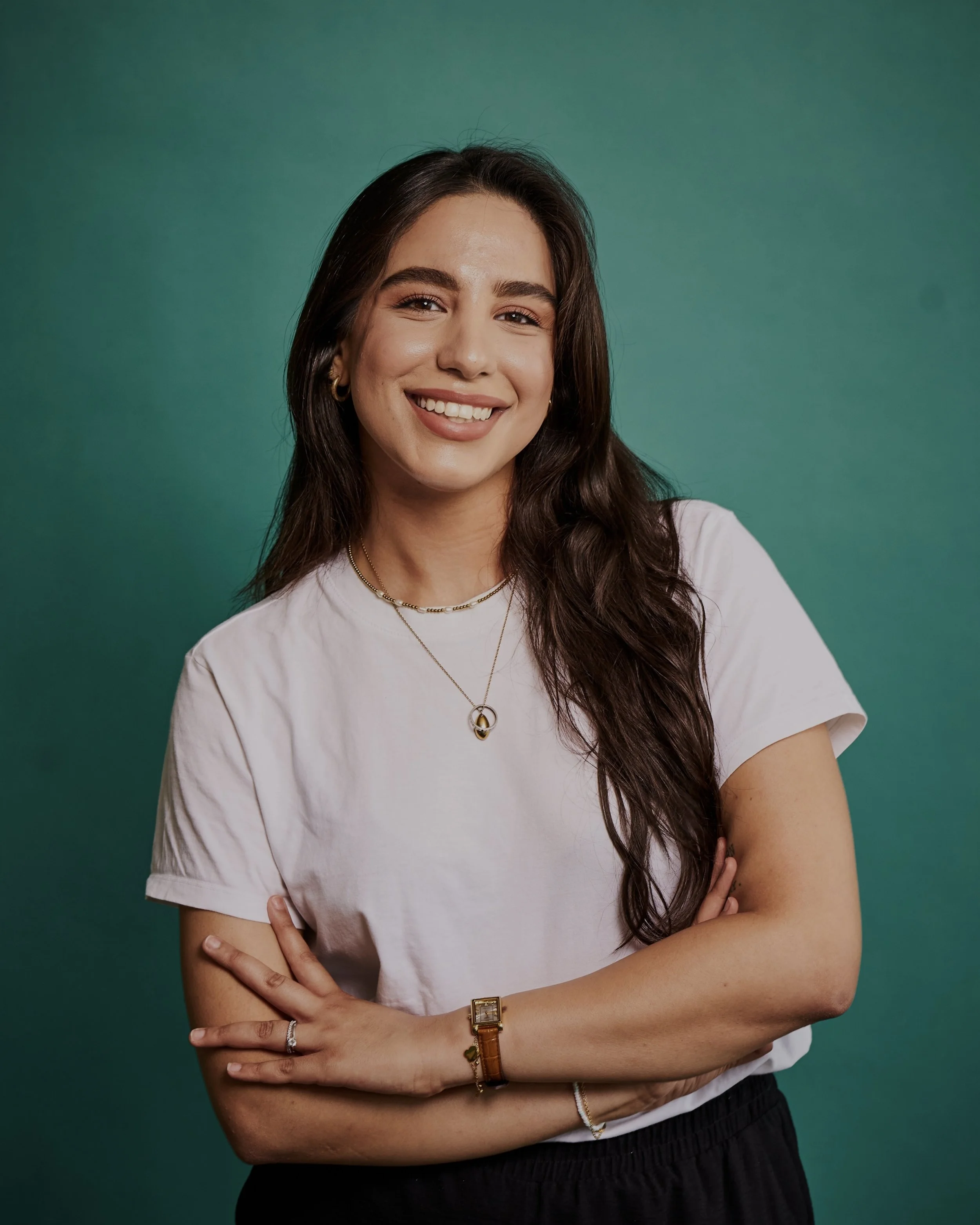 Junge Frau mit dunklen langen Haaren steht vor einem türkisen Hintergrund. Die Arme sind verschränkt vor der Brust und sie lächelt freundlich in die Kamera.
