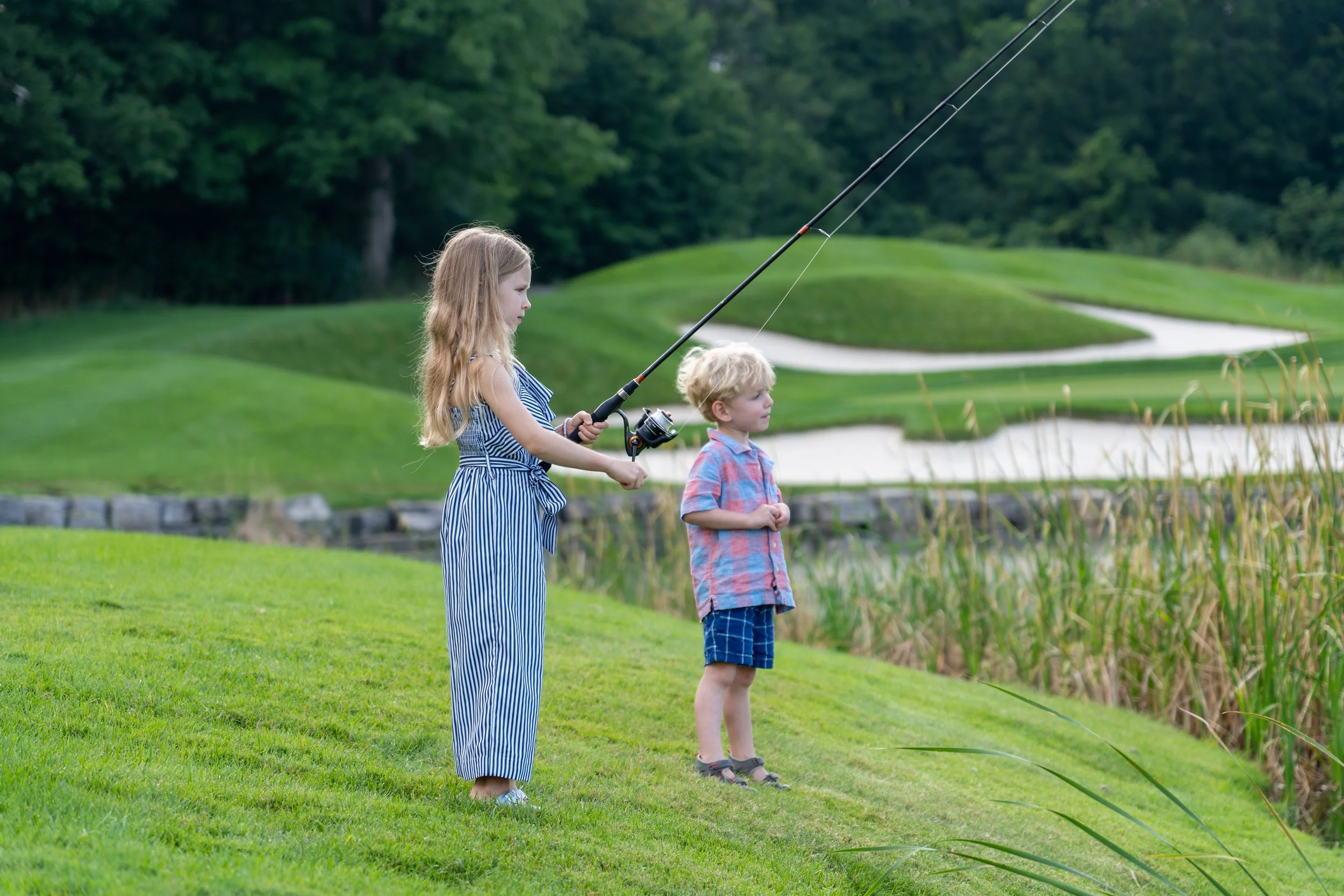 A young girl with long blonde hair wearing a blue and white striped dress fishing with a young boy in a plaid shirt and shorts by a pond in a park or golf course, with green grass, trees, and sand traps in the background.