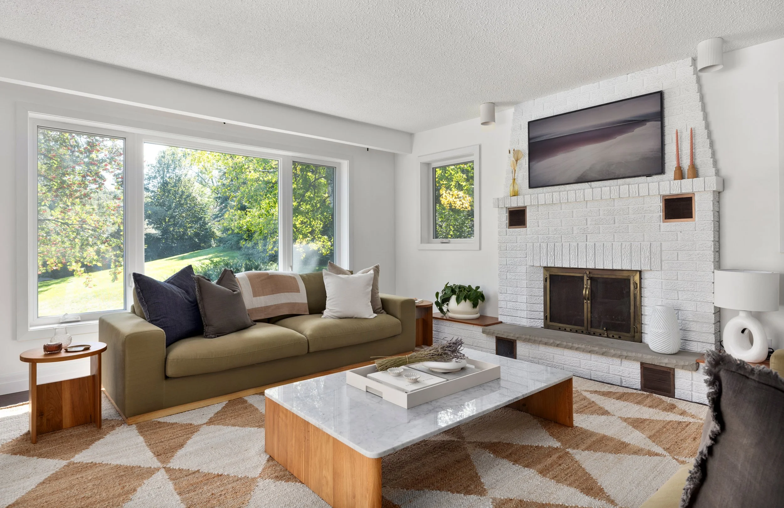 Living room with large window view of greenery, white brick fireplace with a mounted TV, beige sofa with colorful pillows, wooden coffee table with decor, patterned rug, and decorative items on mantel and side table.