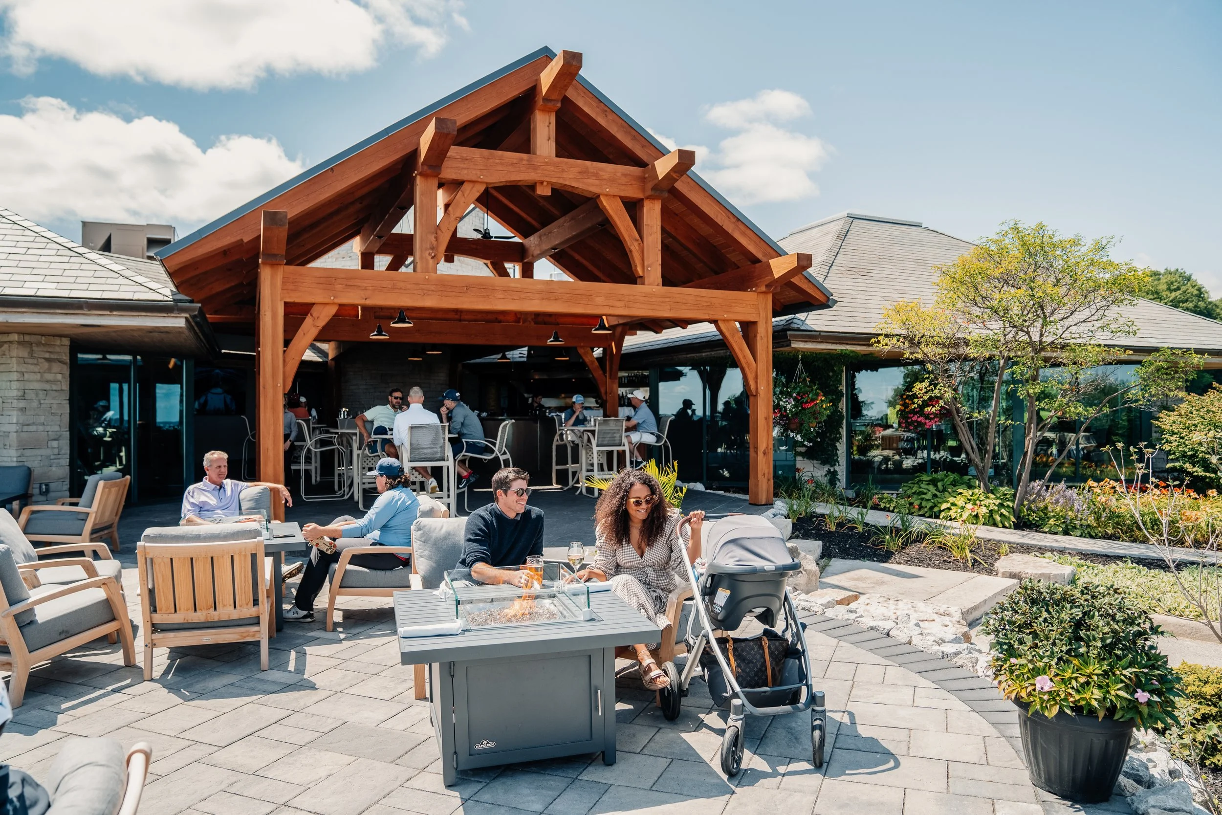 People enjoying outdoor dining area with a barbecue grill, wooden pavilion, and lush greenery.