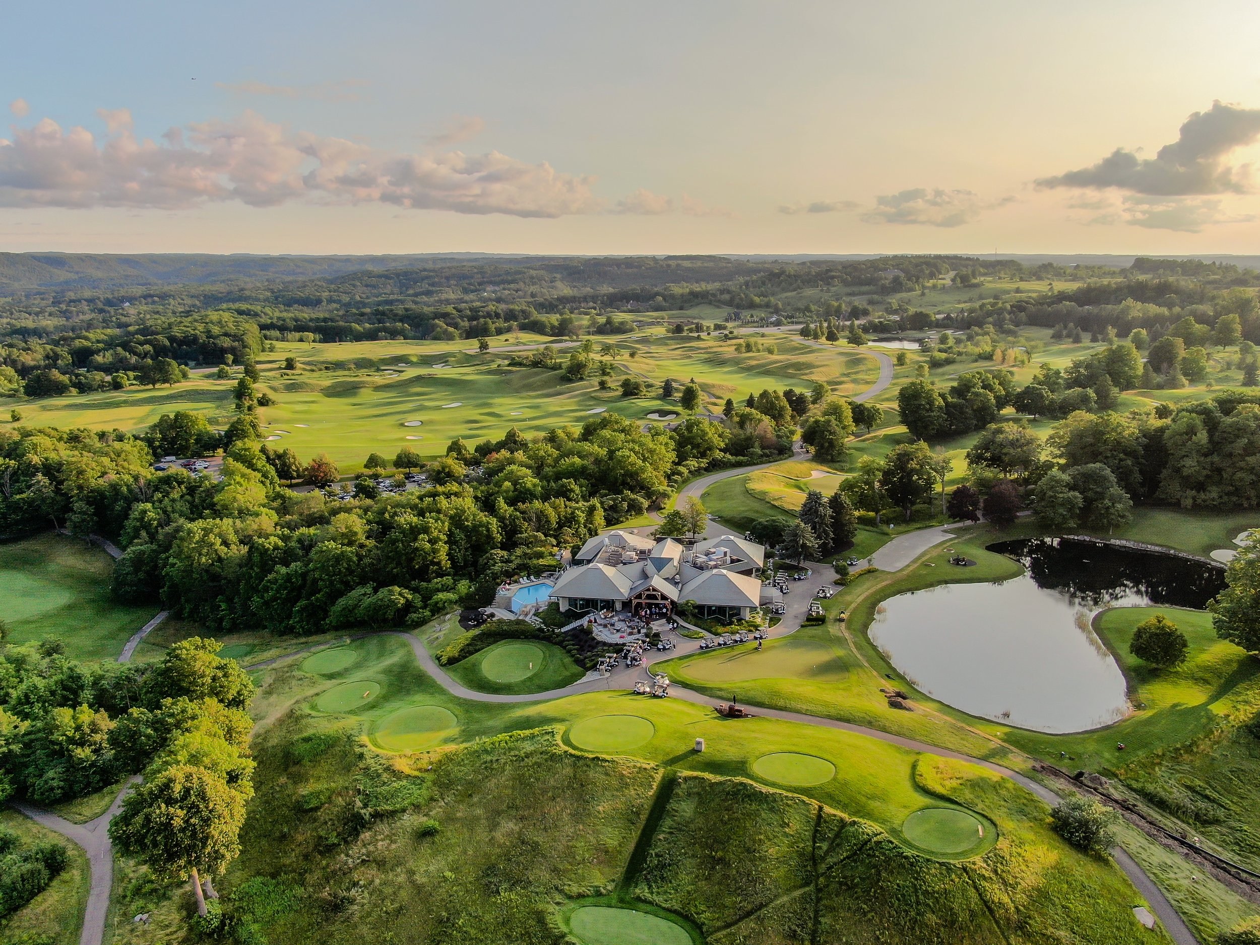 Aerial view of a golf course with green fairways, sand traps, a pond, and a clubhouse surrounded by trees and rolling hills under a partly cloudy sky.