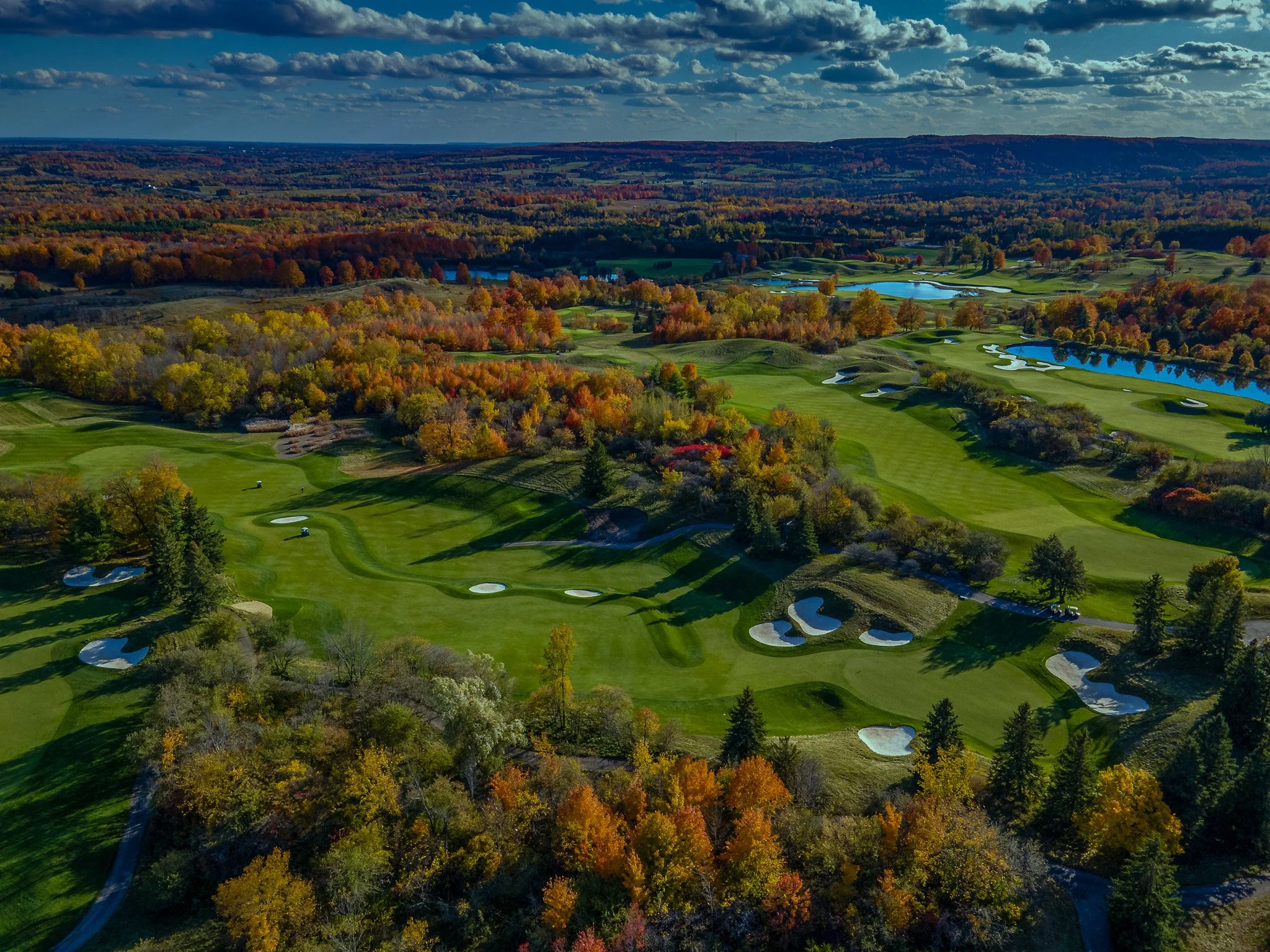 An aerial view of a golf course surrounded by colorful fall trees, with lakes and rolling hills in the distance under a partly cloudy sky.