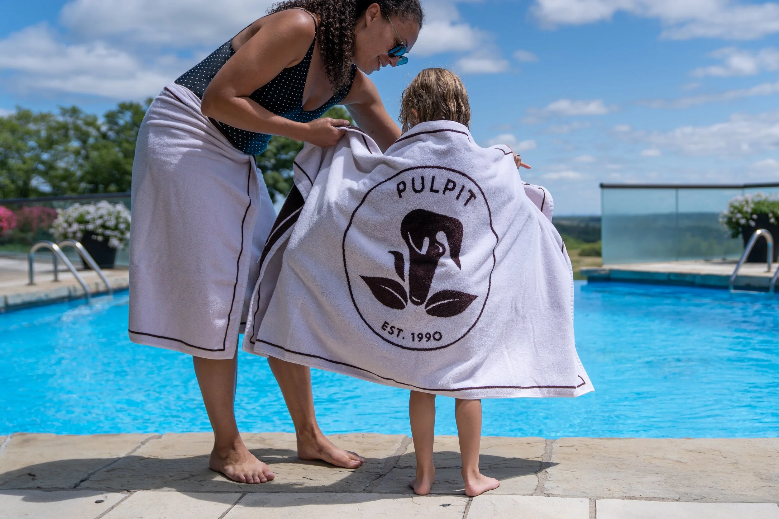 A woman helping a young child with a towel by a swimming pool on a sunny day.
