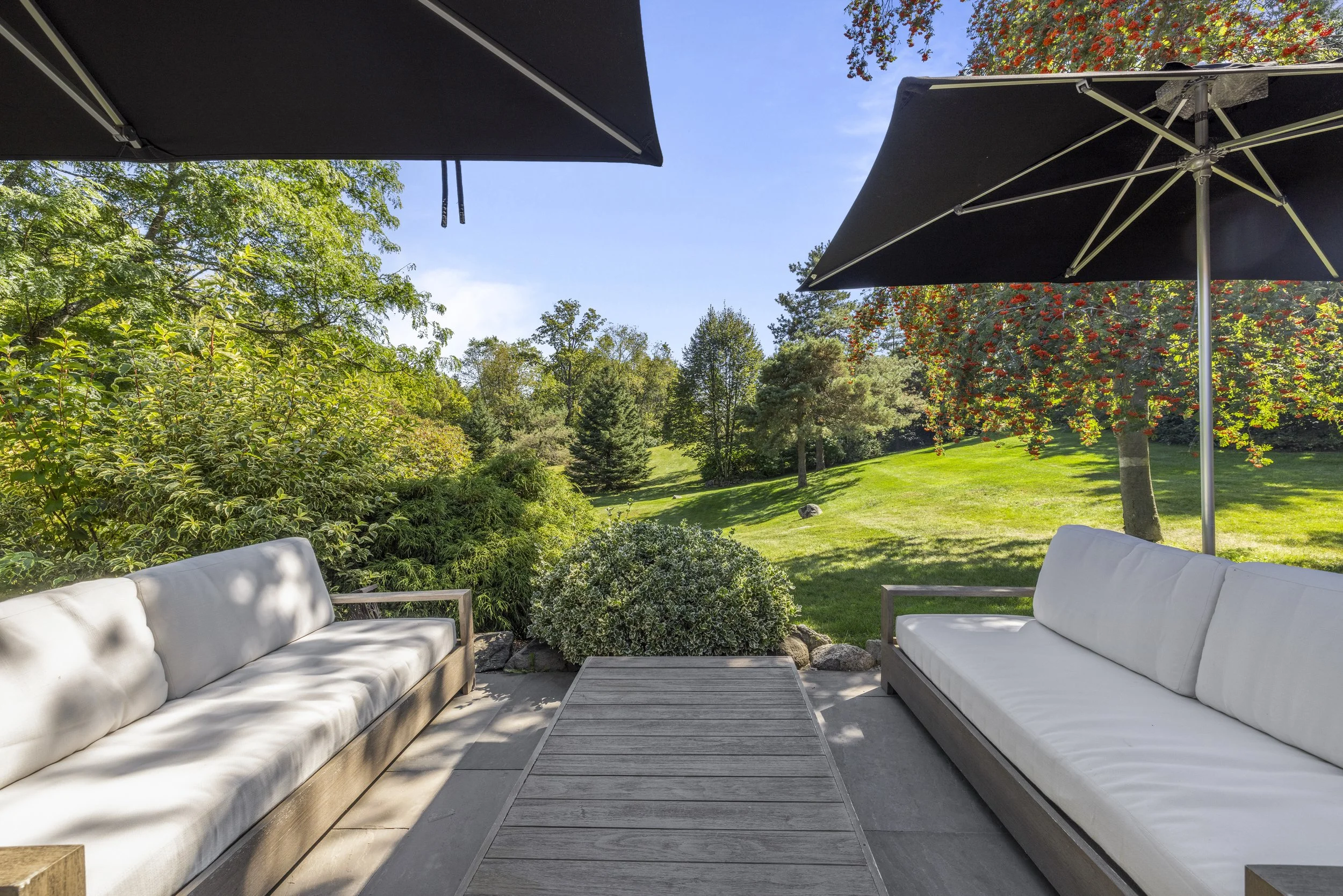Outdoor patio with two white cushioned couches, a wooden table, black umbrellas, and a lush green garden with trees and grass in the background.