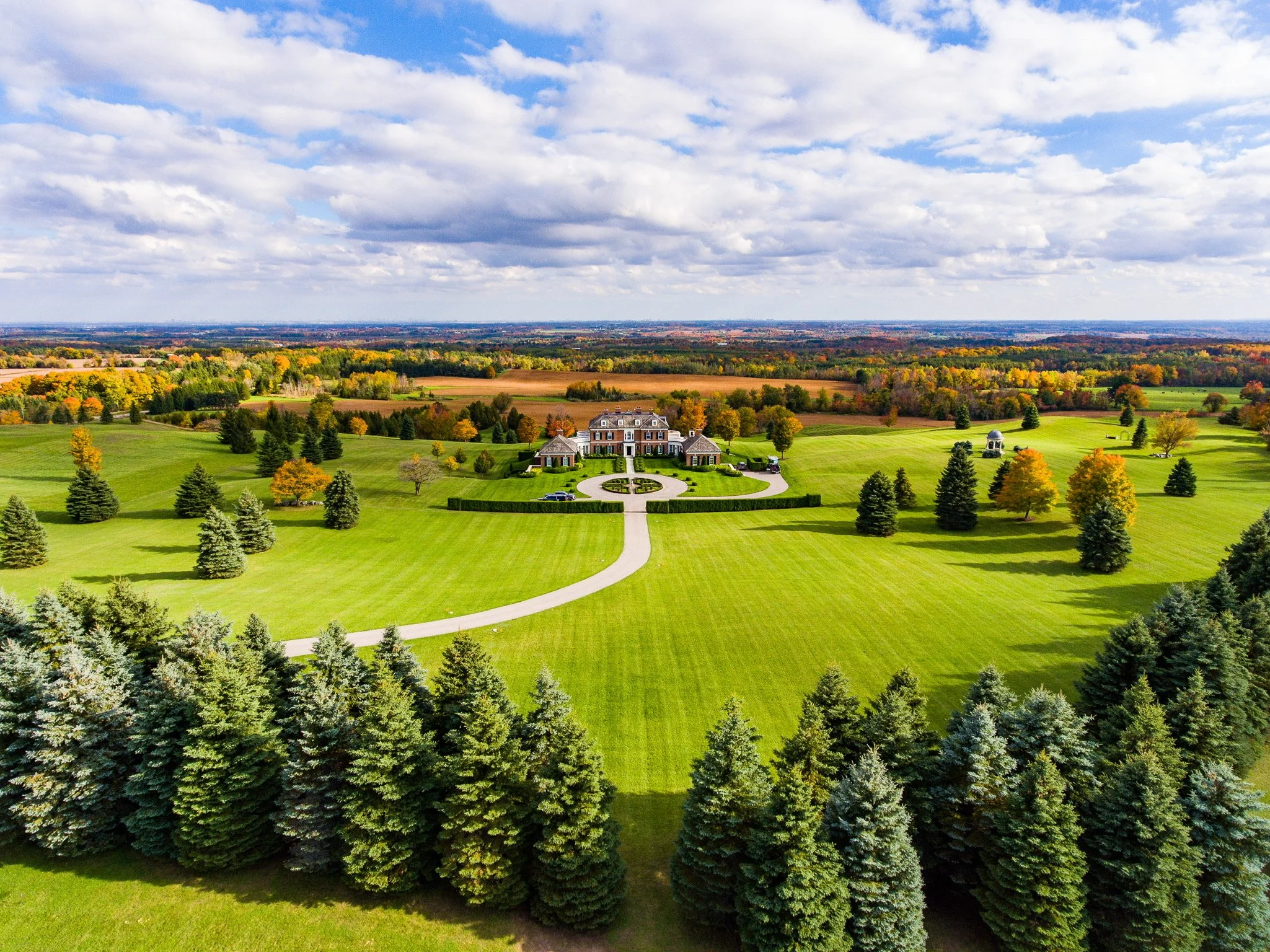 A large house with a circular driveway surrounded by a well-maintained lawn and trees, set in a broad open landscape with fields and distant forests under a partly cloudy sky.