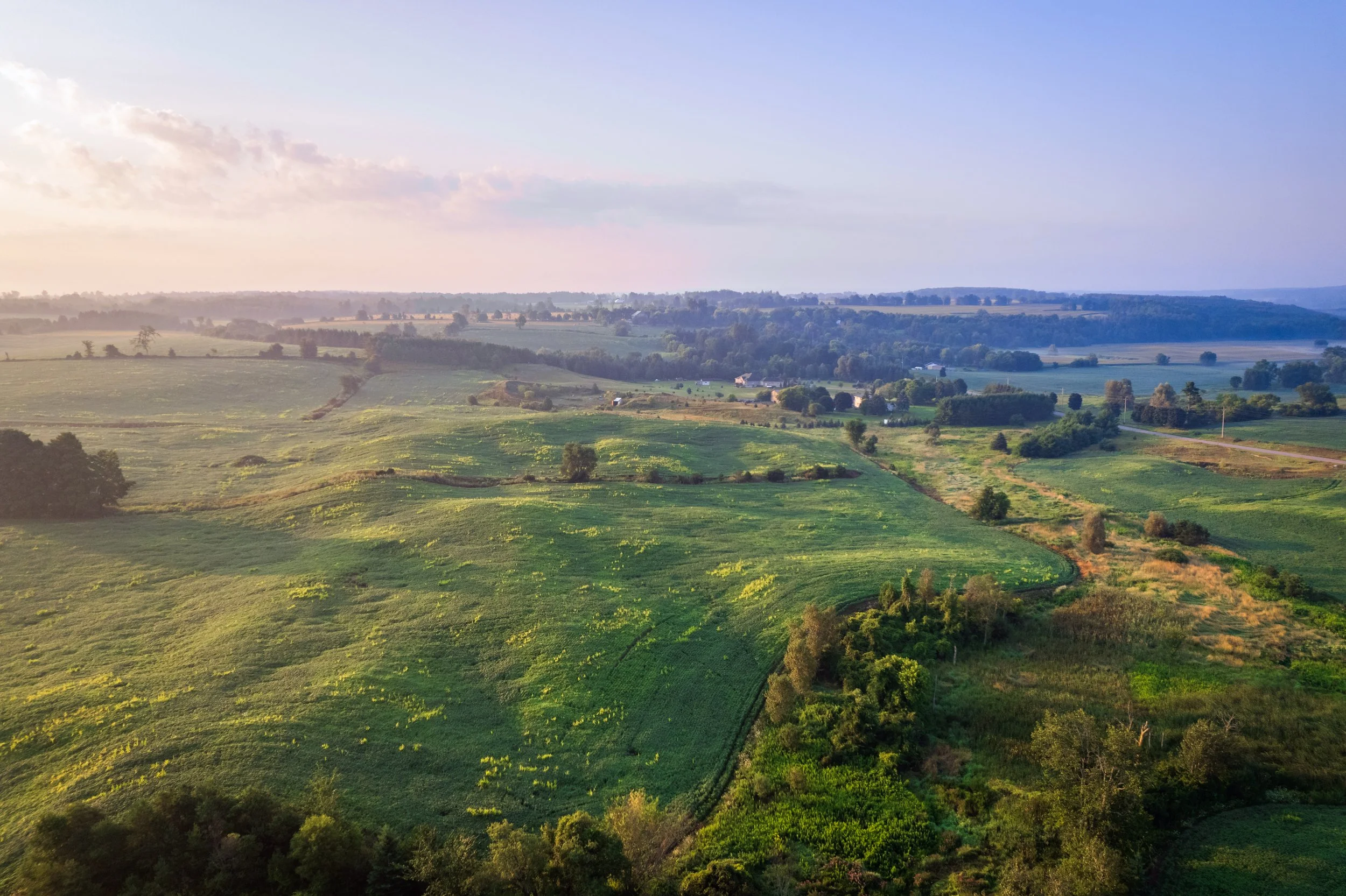 Aerial view of a lush green countryside with rolling hills, small clusters of trees, and open fields under a partly cloudy sky.